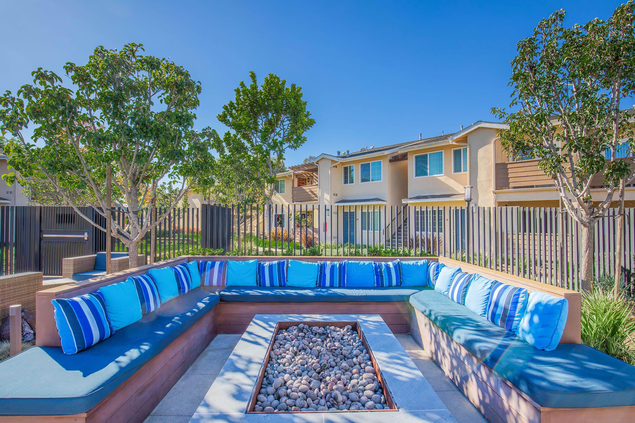 A modern outdoor lounge area featuring a large, curved sofa with blue and striped cushions. In the center, there's a fire pit filled with pebbles. Surrounding the space are trees and a fenced backyard, with nearby apartment buildings in the background under a clear blue sky.