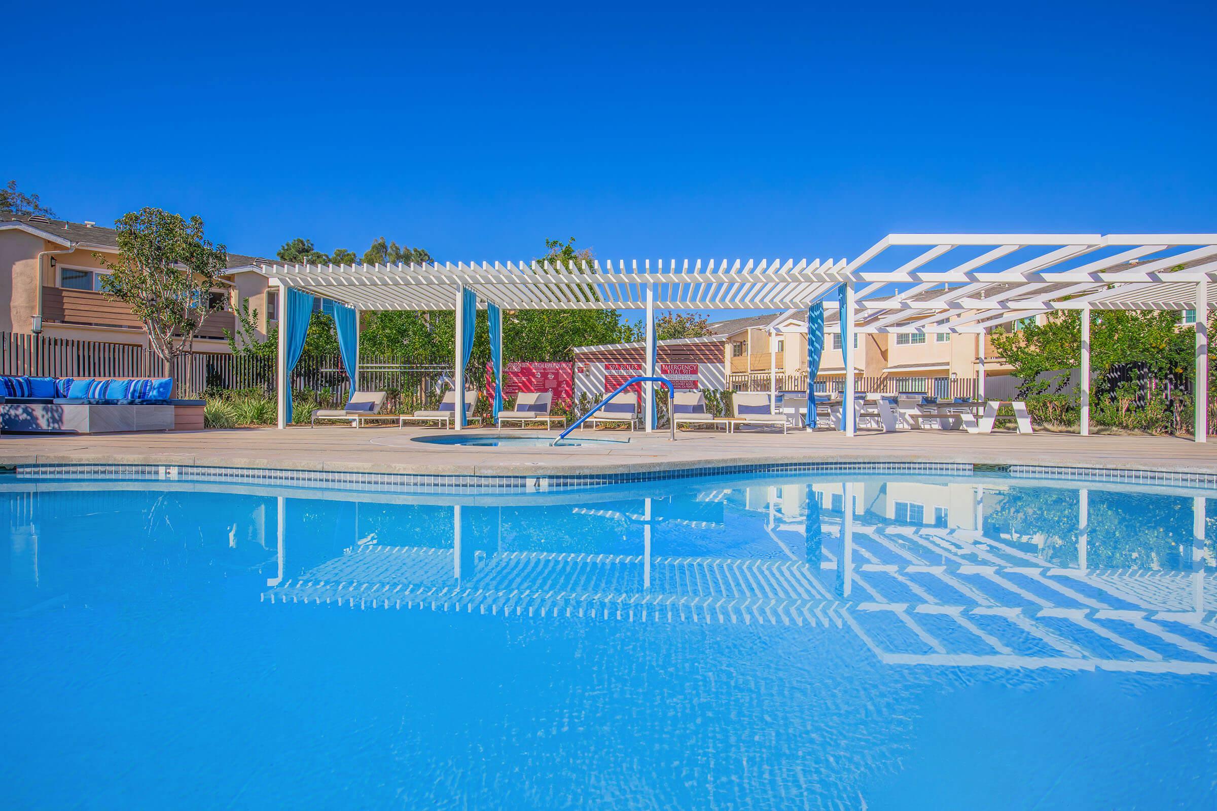 A clear blue swimming pool surrounded by shaded lounge areas and pergolas, with reflections on the water's surface. Neatly arranged chairs and tables provide a relaxing outdoor space in a residential setting. Bright, sunny sky adds to the inviting atmosphere.