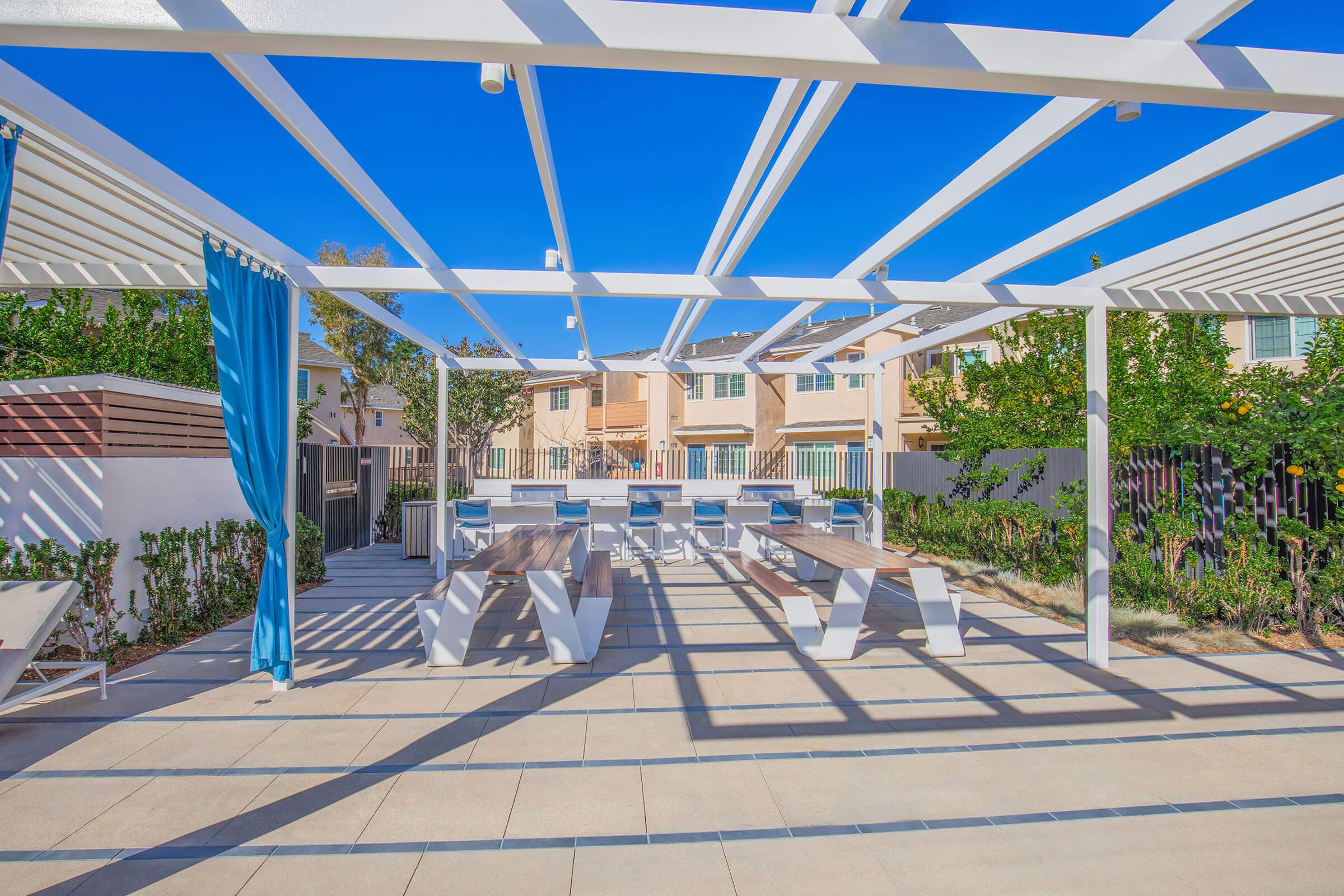 Outdoor space featuring a pergola with blue curtains, surrounded by trees and shrubs. There are long white tables and benches for seating. In the background, residential buildings are visible under a clear blue sky. Sunlight casts shadows on the paved ground, creating a lively atmosphere.