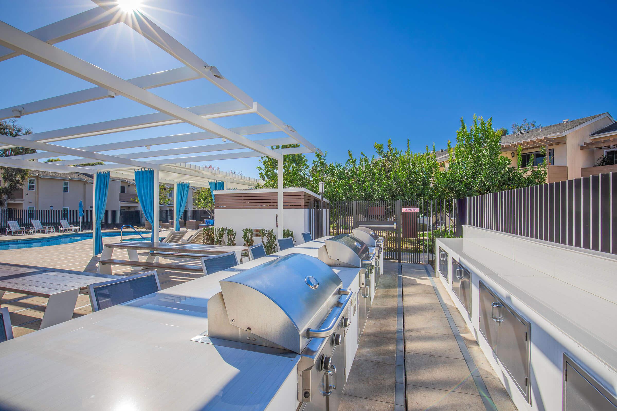 Outdoor barbecue area featuring multiple stainless steel grills under a white pergola, with a swimming pool and lounge chairs in the background. Lush greenery is visible beyond a fenced area, and the sky is clear and sunny.