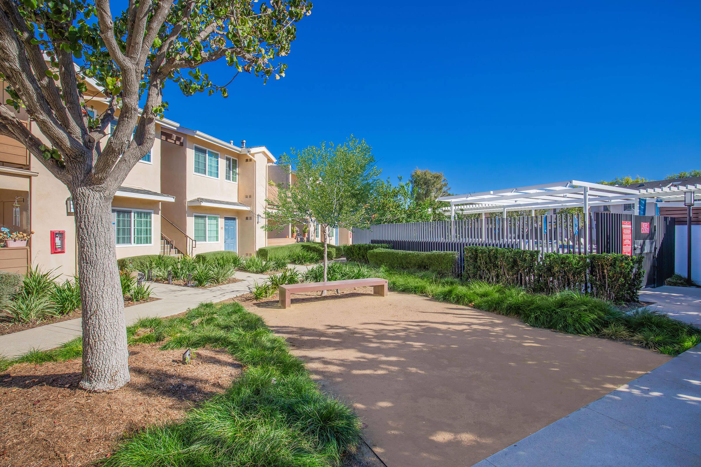 A landscaped outdoor area with a sandy space and a wooden bench surrounded by grass. In the background, there are two buildings with several windows and a clear blue sky. The area is well-maintained with trees and shrubs, creating a pleasant and inviting atmosphere.
