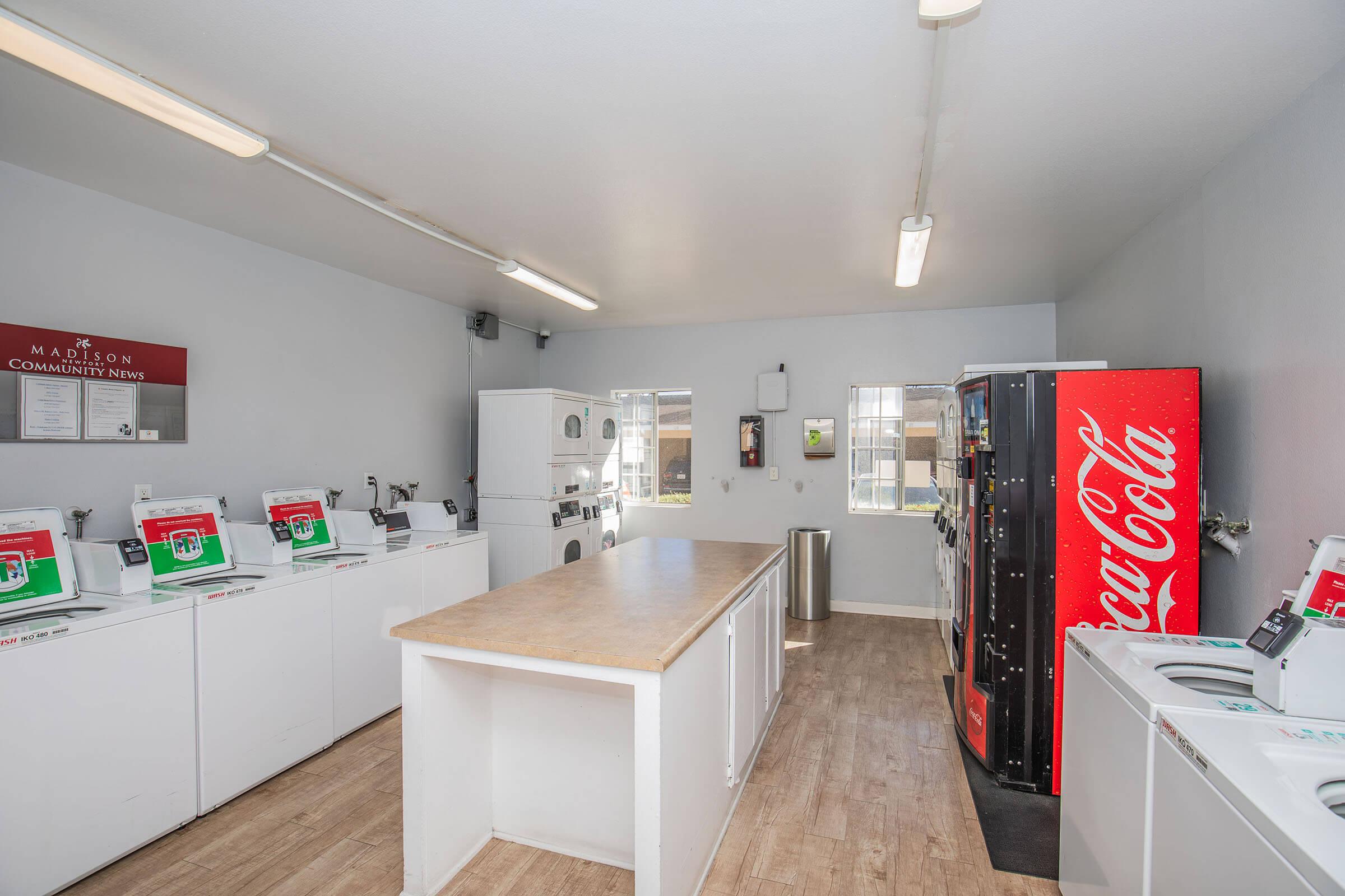 A clean laundry room featuring multiple washing machines and dryers arranged along a countertop. There's a Coca-Cola vending machine on one side, and large windows provide natural light. The walls are painted light gray, creating a bright and inviting atmosphere.