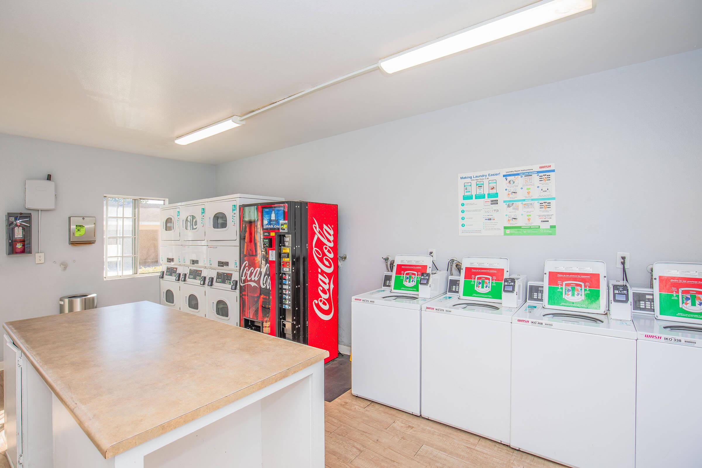 Interior view of a laundry room featuring several washing machines and a vending machine stocked with Coca-Cola products. The walls are light-colored, with a window providing natural light, and there are informational posters about laundry usage.