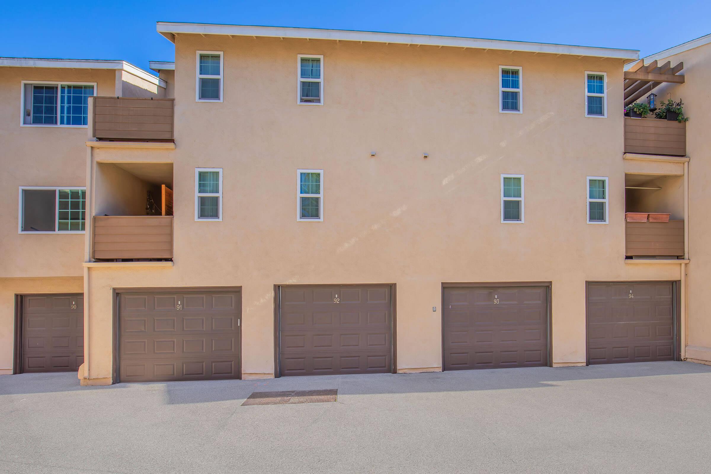 A tan multi-level building with multiple garage doors at the bottom. The upper levels feature several windows and balconies. The structure is set against a clear blue sky, and the ground is paved.