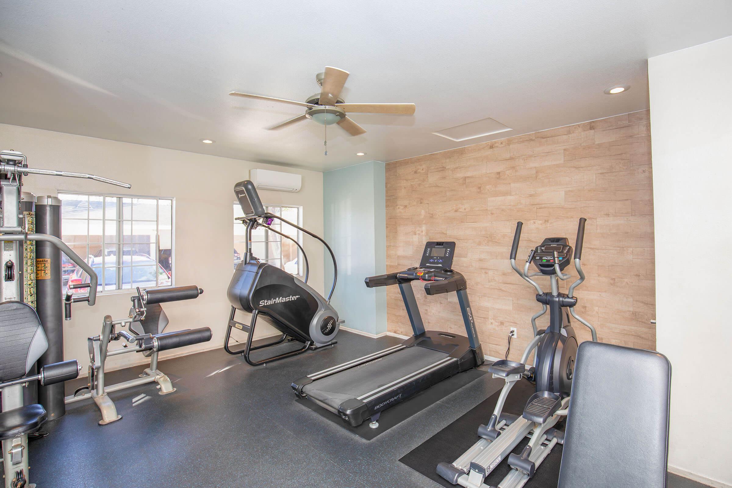 A modern gym interior featuring various fitness equipment, including a treadmill, elliptical machine, and weight machines. The space has wood paneling on one wall, a ceiling fan, and bright lighting, creating a welcoming atmosphere for workouts. Large windows allow natural light to enter.