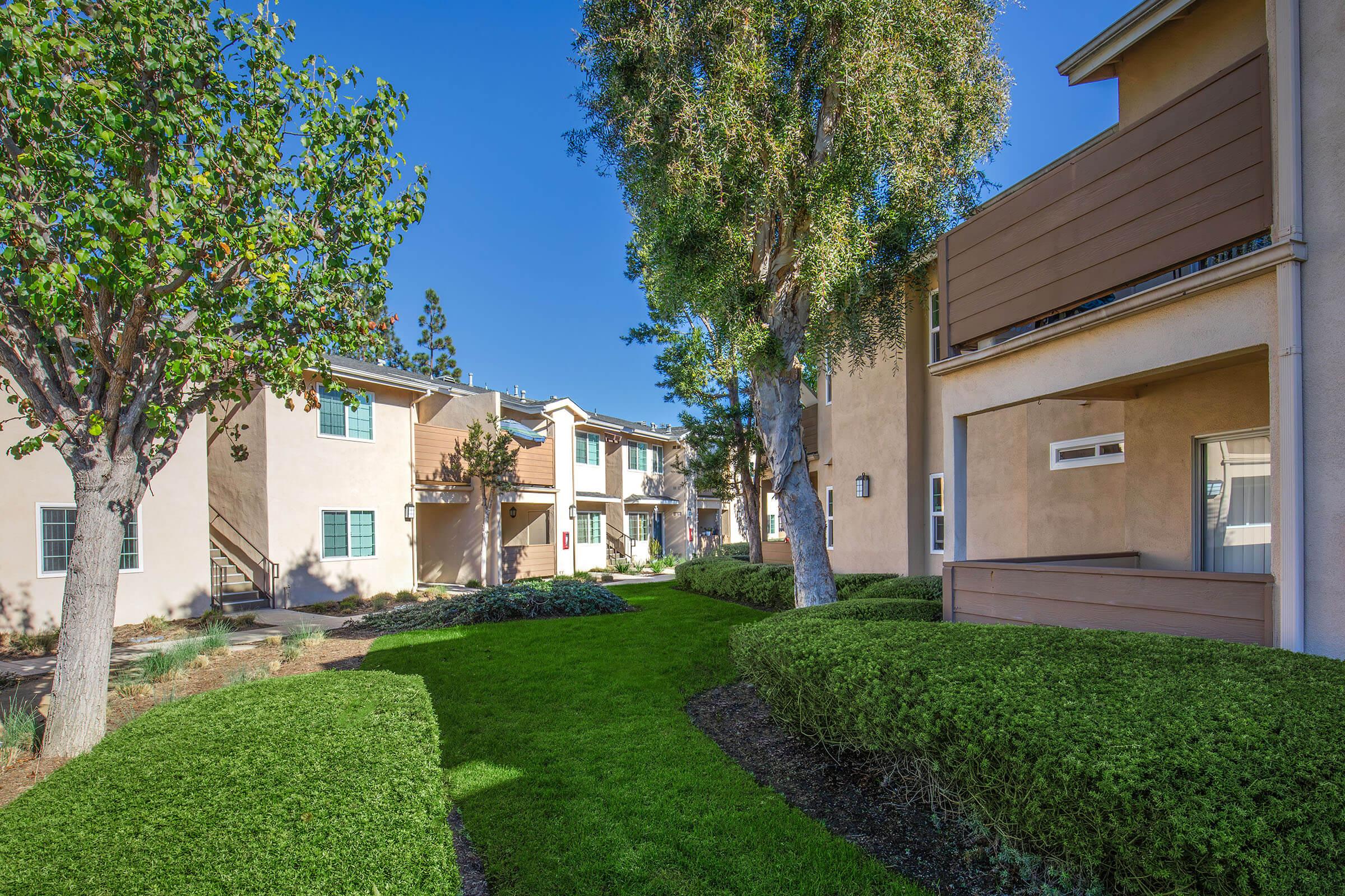 A well-maintained residential area featuring green lawns and landscaped shrubs. There are two-story buildings with balconies and windows, surrounded by trees under a clear blue sky, suggesting a tranquil and inviting living environment.