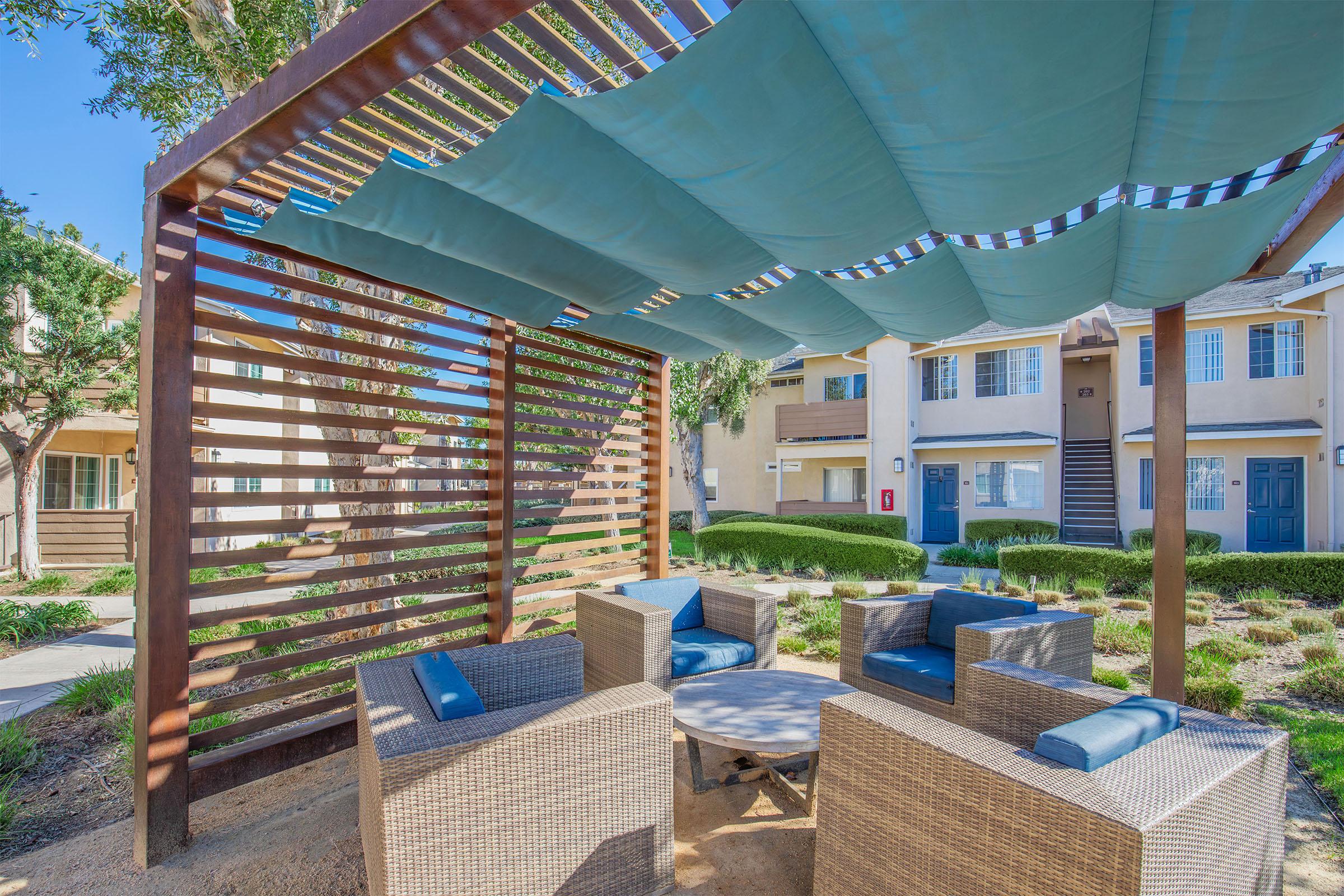A cozy outdoor seating area featuring modern wicker chairs and a round table, shaded by blue fabric drapes. The surrounding landscape includes green plants and a well-maintained walkway, with residential buildings visible in the background under a clear blue sky.