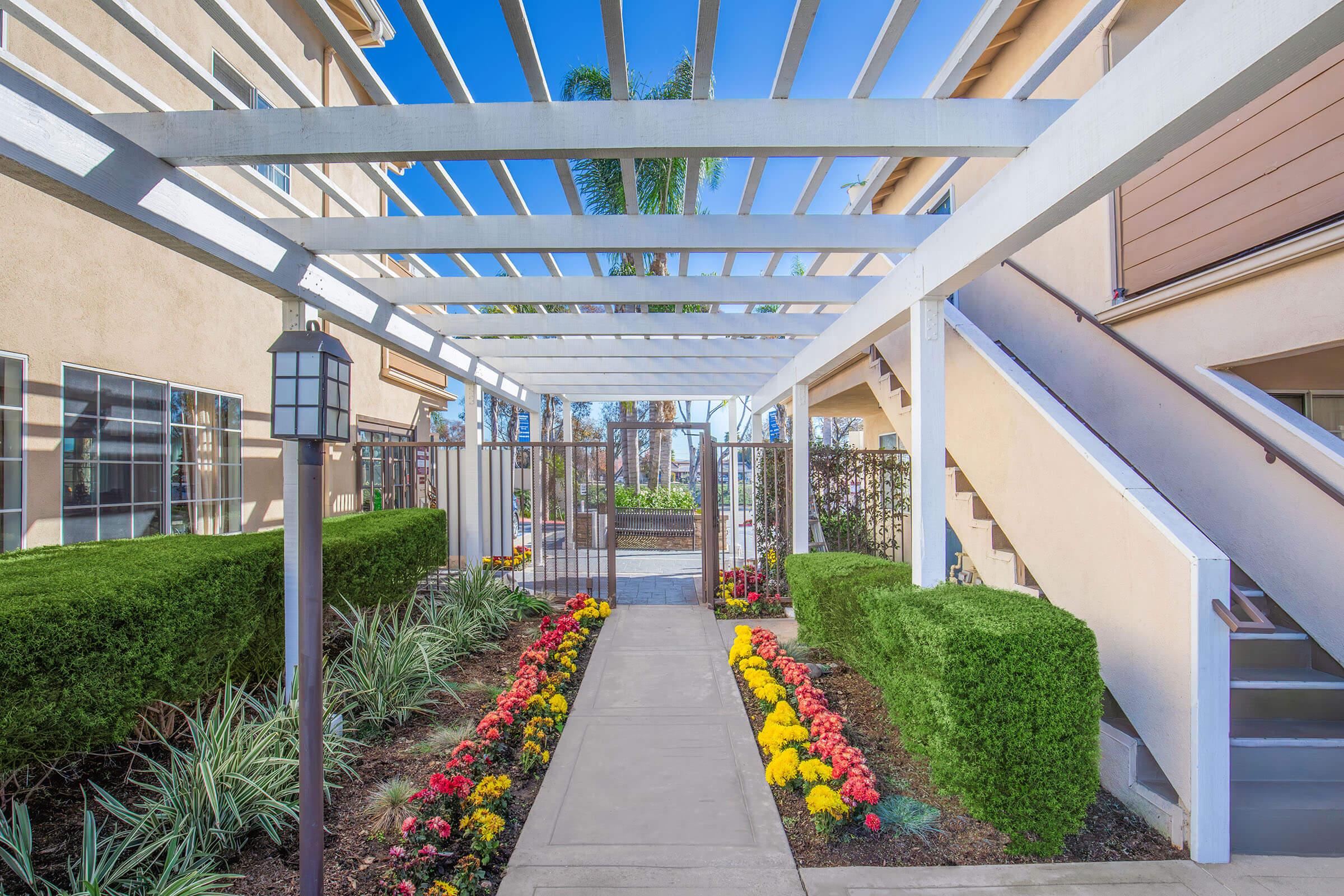 A landscaped pathway leads through a white trellis structure, flanked by colorful flower beds in bloom. On either side, lush green hedges are neatly trimmed. The background features modern building architecture under a clear blue sky, enhancing the inviting atmosphere of the outdoor space.