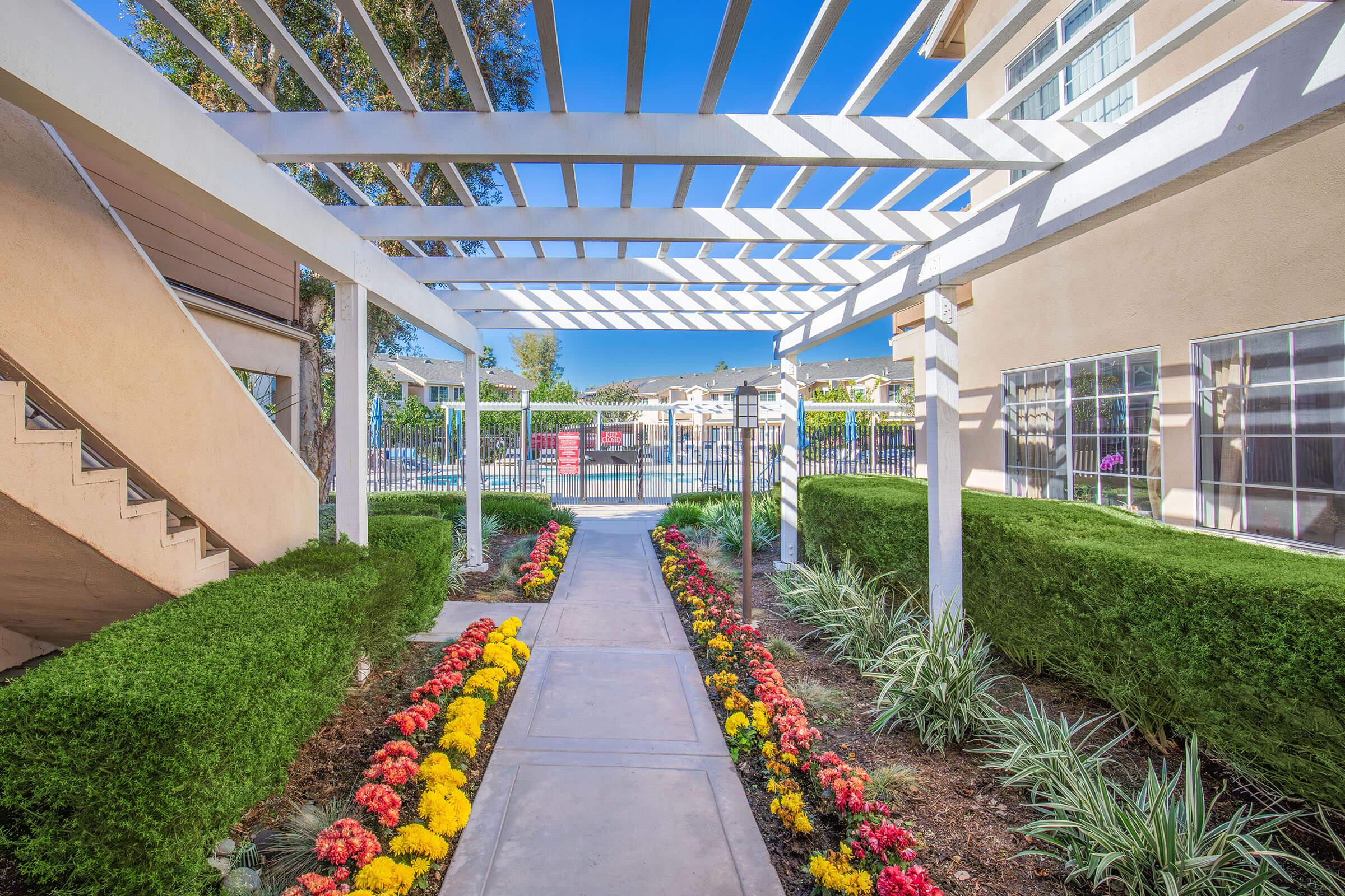 A landscaped walkway bordered by colorful flowers leads through a white pergola structure. On one side, there are neatly trimmed hedges and stairs. In the background, a pool area is visible, enclosed by a fence, under a clear blue sky.