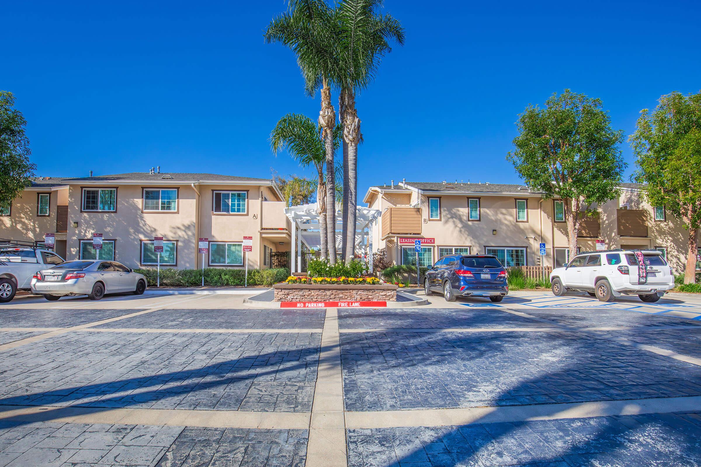 A view of a residential complex featuring two buildings surrounded by palm trees and vibrant landscaping. Several parked cars are visible, and the sky is clear blue, indicating a sunny day. The entrance includes a decorative sign and flower beds, creating an inviting atmosphere.