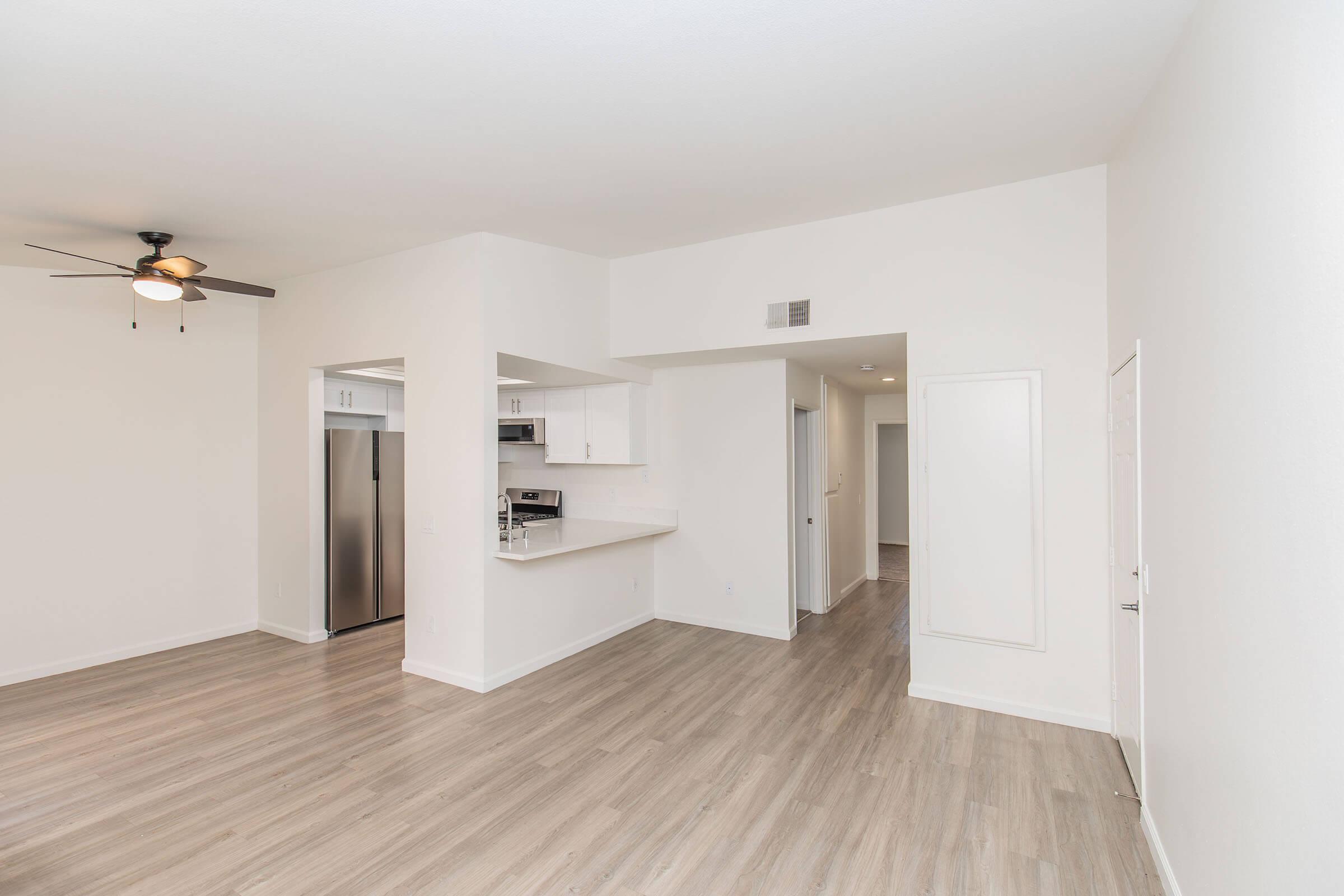 A spacious, modern living area featuring light-colored walls and a wooden floor. To the left, there's a kitchen with a countertop and stainless steel appliances, including a refrigerator. The room is bright with a ceiling fan and open space leading to other areas. The overall aesthetic is clean and minimalist.