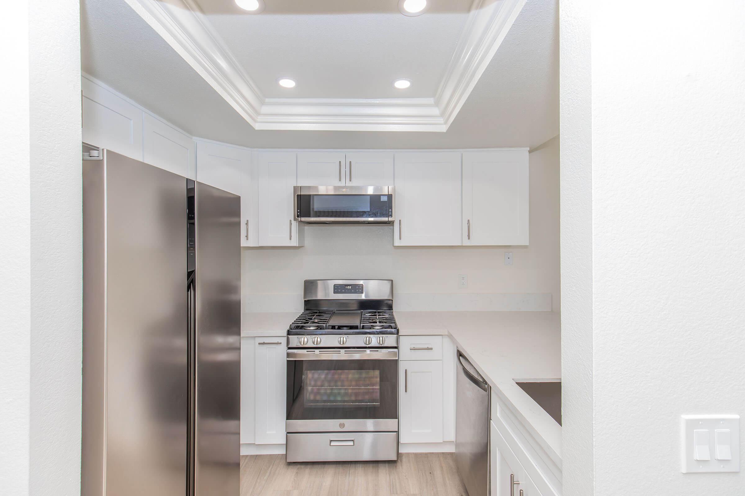 A photo of a modern kitchen featuring a stainless steel refrigerator, a gas range with an overhead microwave, and white cabinetry. The space is well-lit with recessed ceiling lights and has a clean, contemporary design with light-colored countertops.