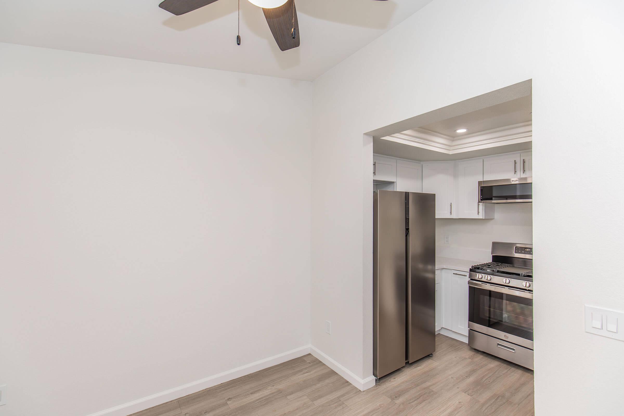A modern kitchen featuring stainless steel appliances, including a refrigerator and stove. The kitchen has white cabinetry and light-colored walls, with a ceiling fan visible. The flooring is a light wood tone, creating a clean and spacious look.