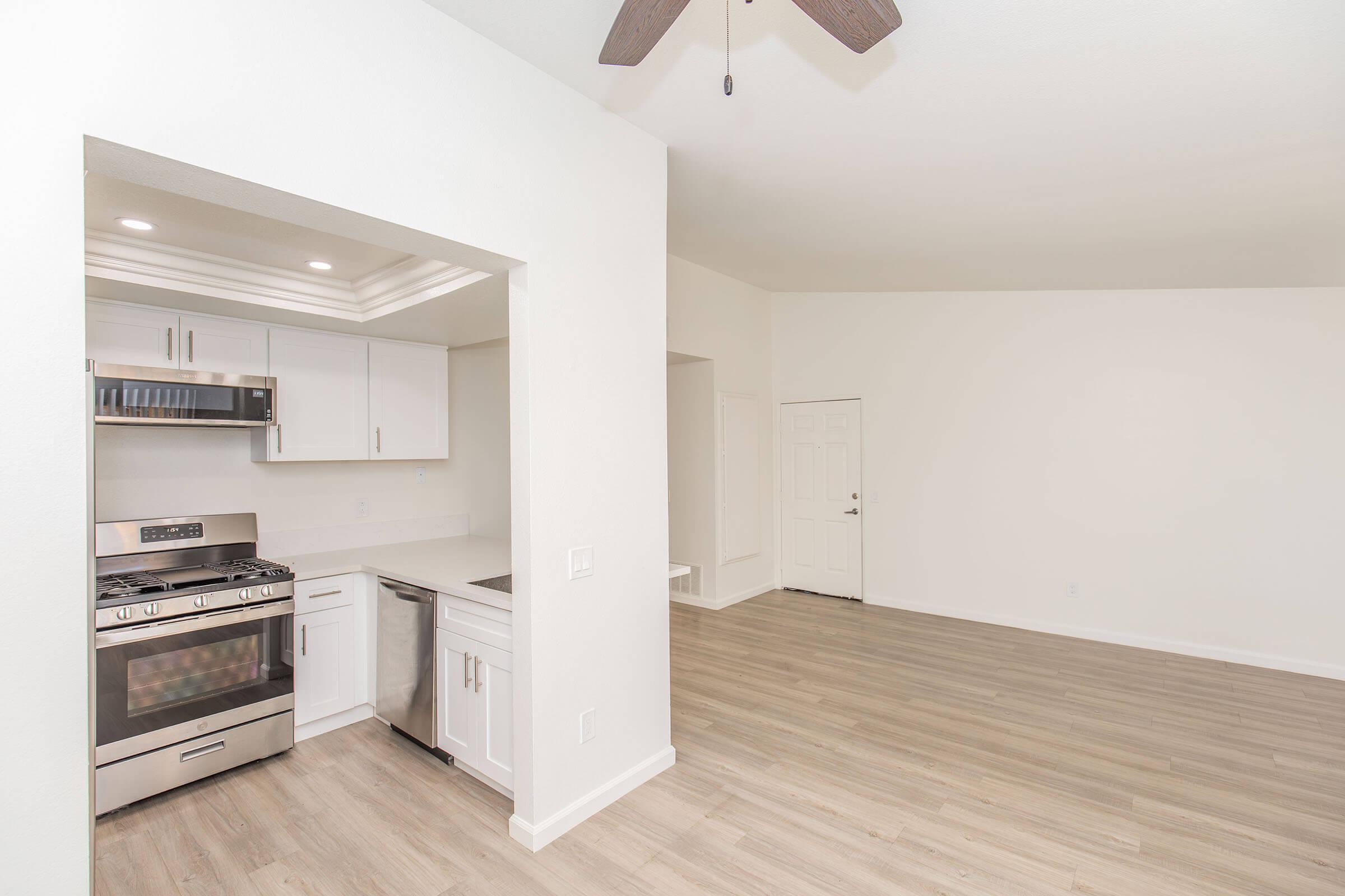 Modern kitchen with stainless steel appliances, including a gas stove and microwave, adjacent to a spacious living area featuring light wood flooring and white walls. A ceiling fan is visible, and there is a doorway leading to another room.