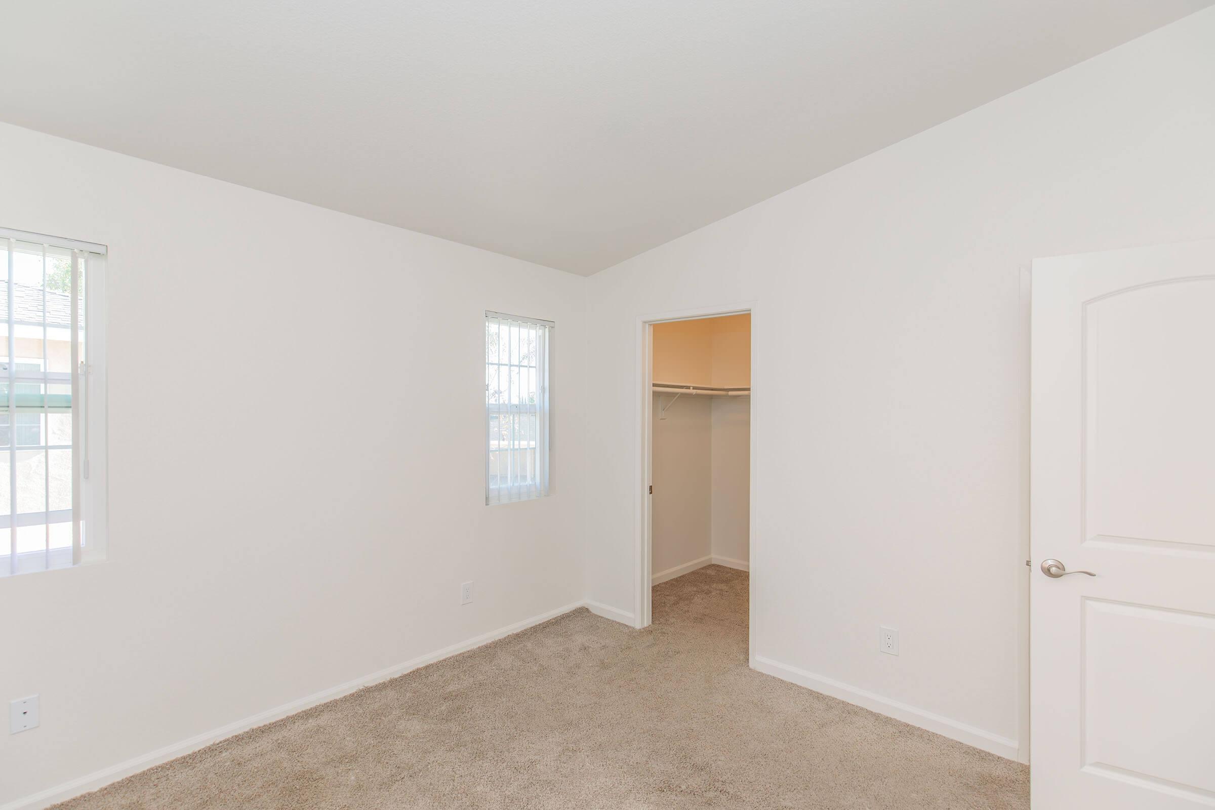 Empty room with light-colored walls and carpeted flooring. Two windows allow natural light, creating a bright atmosphere. A doorway leads to a small closet with an open design. The space is simple and uncluttered, ideal for various decorating options.