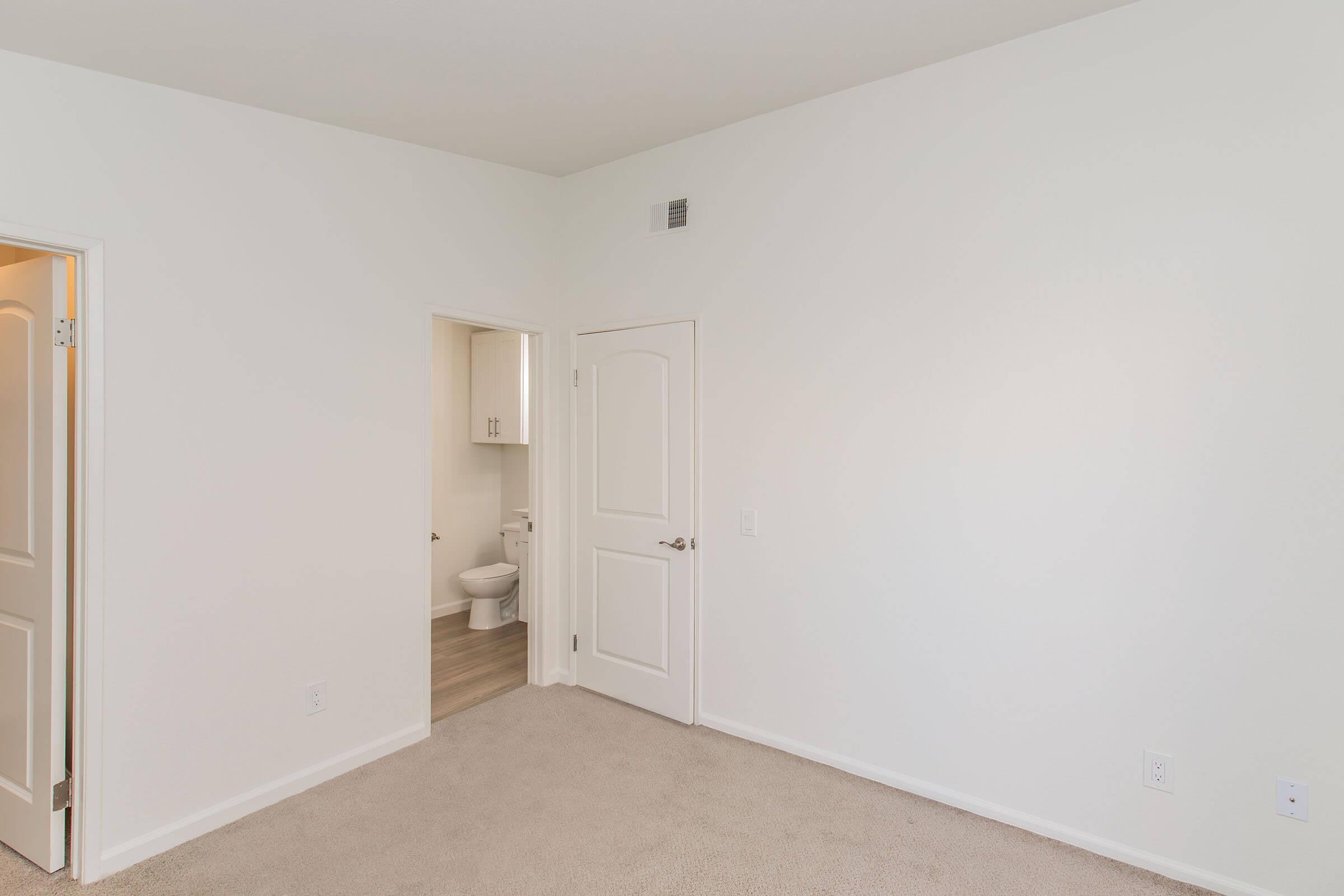 A vacant room with light beige carpet and white walls. There's an open doorway leading to a bathroom with a toilet visible. The space is bright and minimalistic, emphasizing simplicity and neutrality in design.