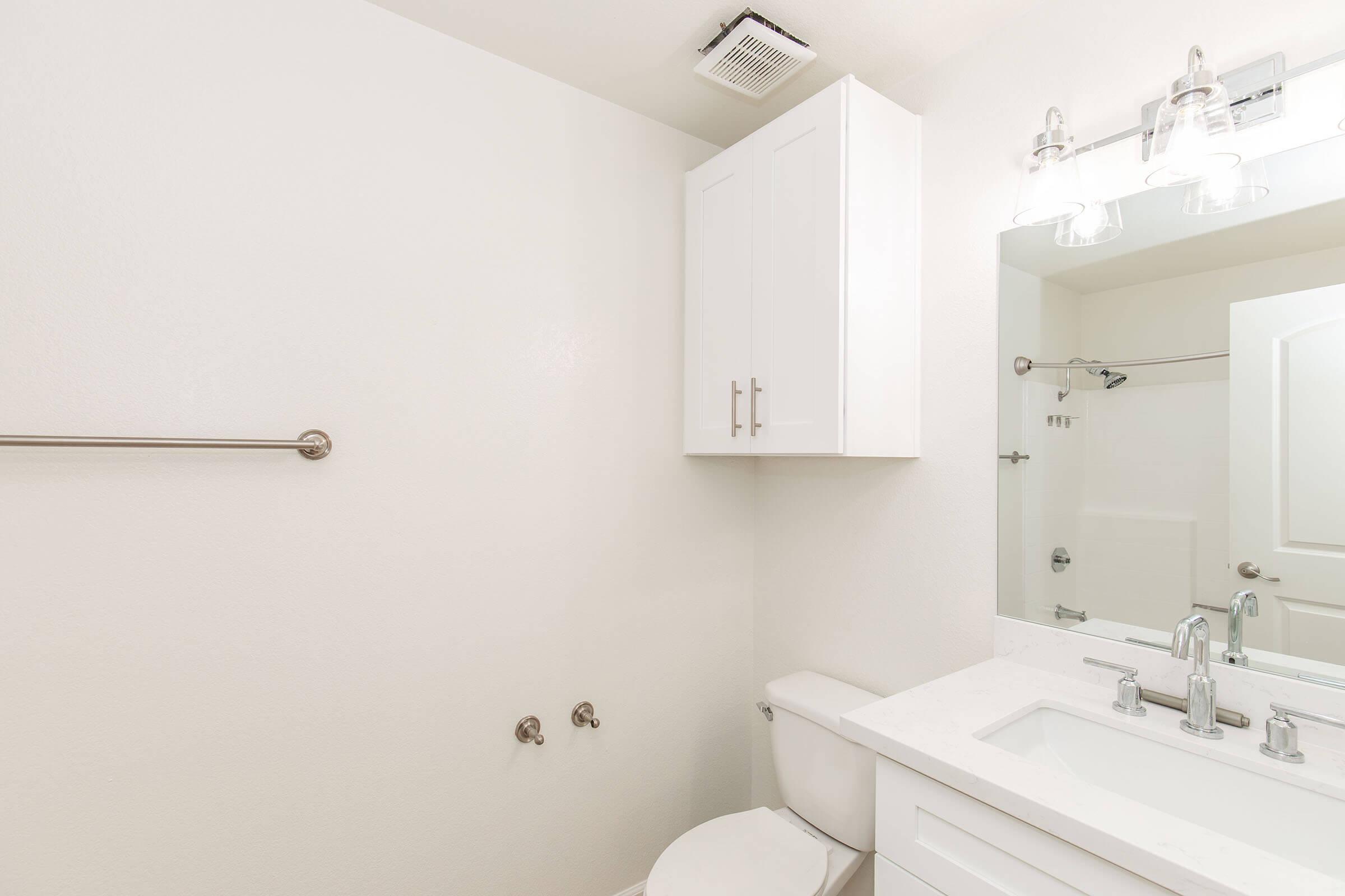A clean, modern bathroom featuring white walls, a white vanity with a sink, a large mirror with light fixtures above, a toilet, and a towel bar. The space is well-lit and minimalistic, emphasizing a fresh and airy atmosphere.