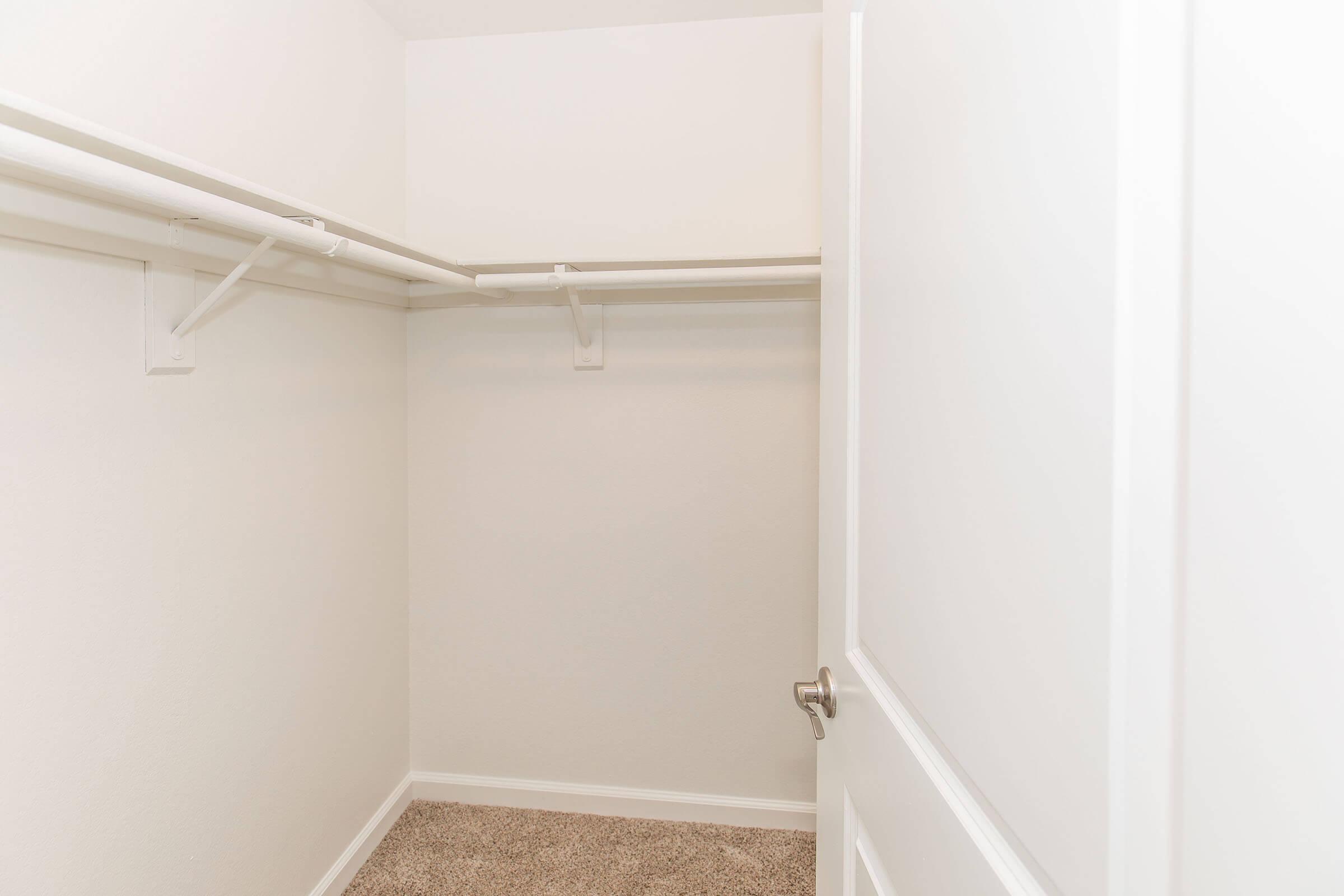 An empty closet with white walls and a light carpet, featuring a hanging rod and shelving. The closet door is partially open.