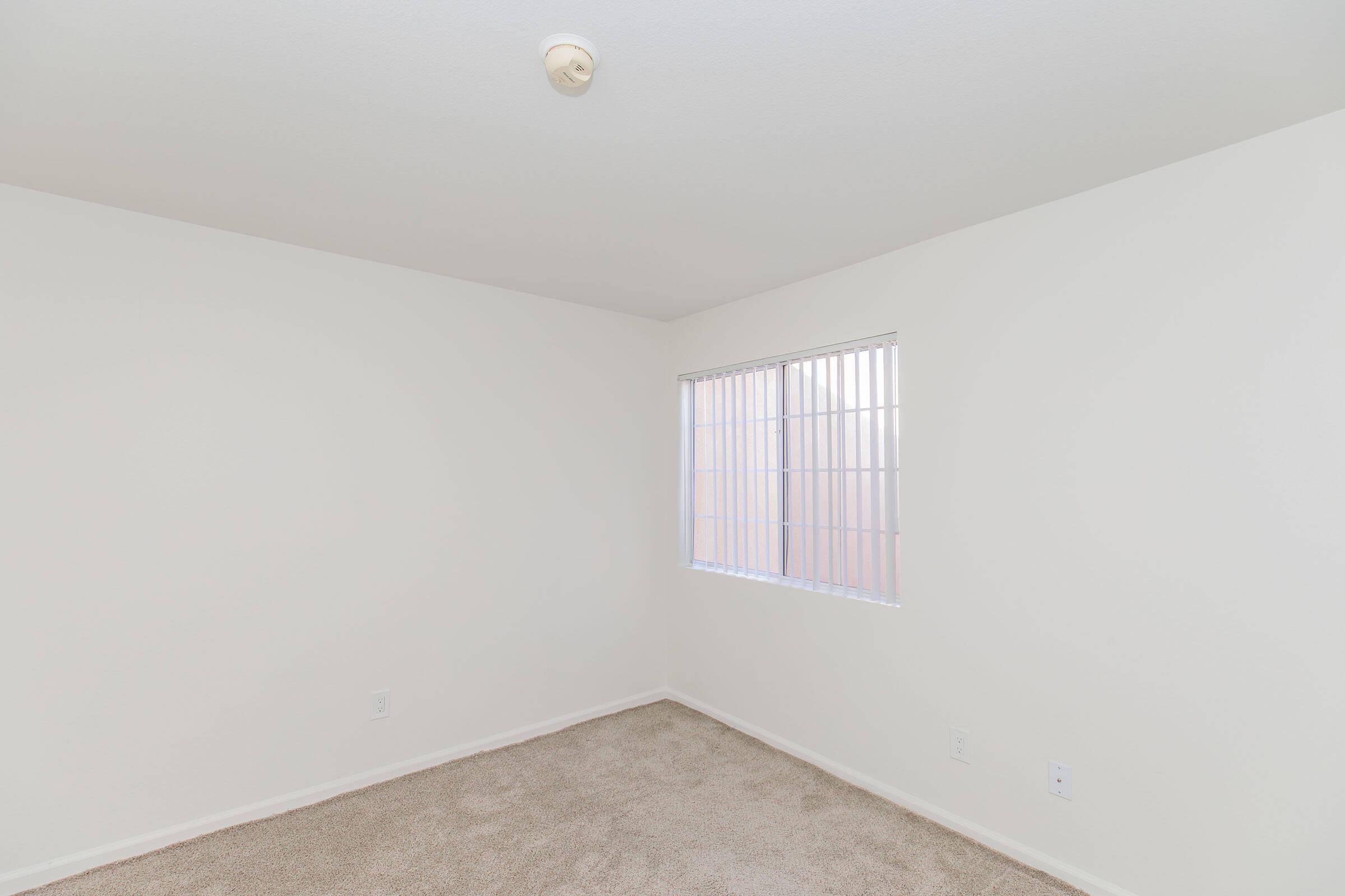 A blank room featuring white walls, beige carpet, and a small window with horizontal blinds, allowing natural light to enter. The ceiling has a light fixture, and the overall space is empty, providing a clean and minimalist look.