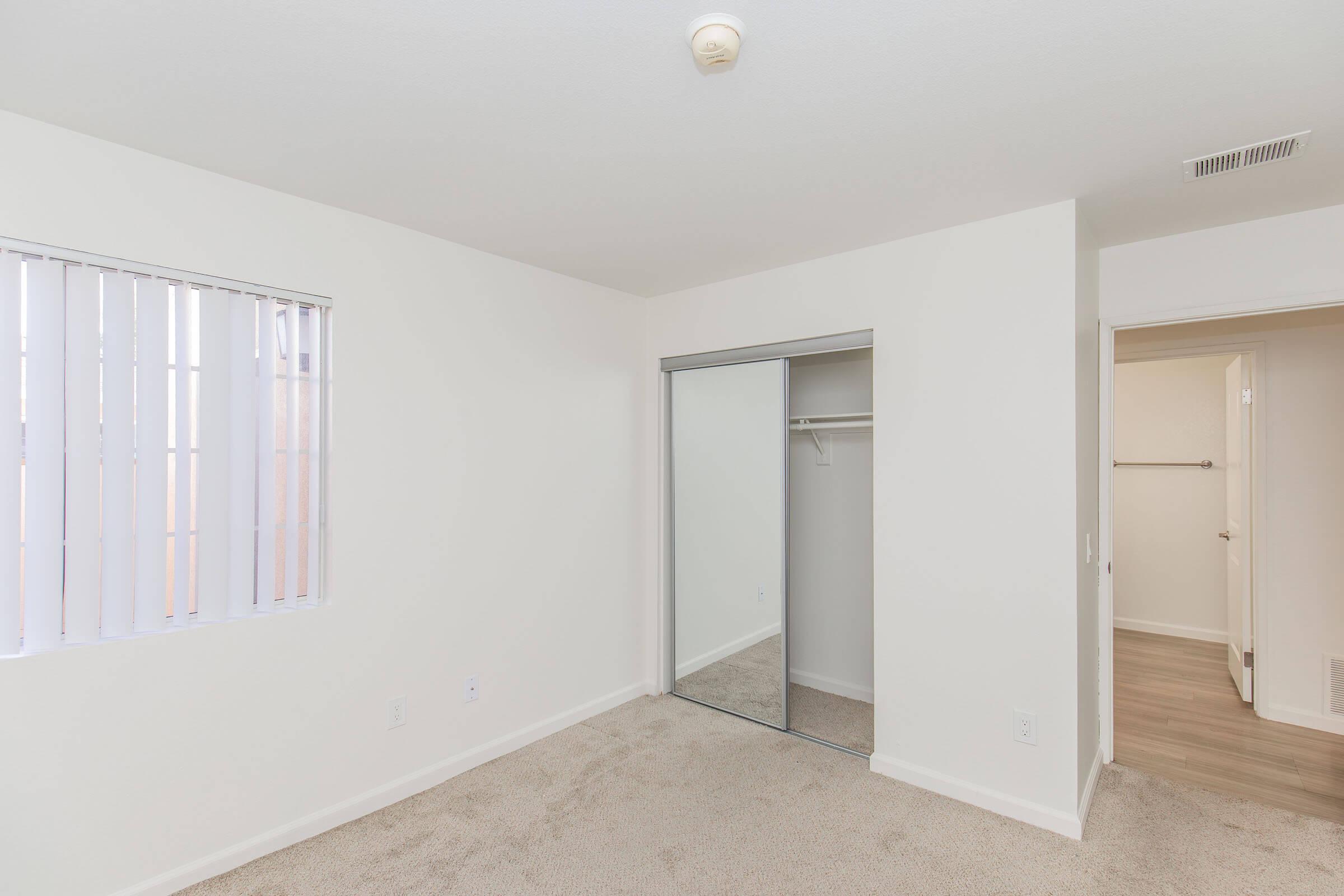 A vacant bedroom featuring light-colored walls, beige carpet, and a large window with blinds. There is a mirrored closet door on the left side and a doorway leading to another room on the right. The overall space appears clean, bright, and ready for occupancy.