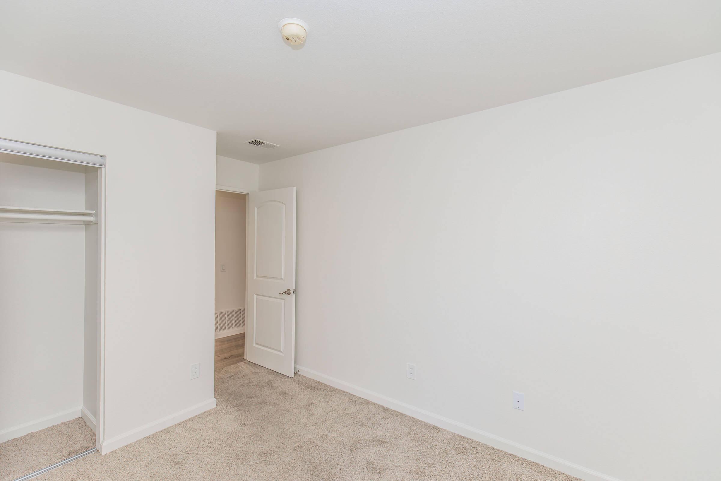 A clean, empty bedroom featuring beige carpet, white walls, and a small closet on the left. There's a doorway leading to another room in the background, and overhead lighting is provided by a simple ceiling fixture. The space is well-lit and unadorned, suitable for personal decoration.