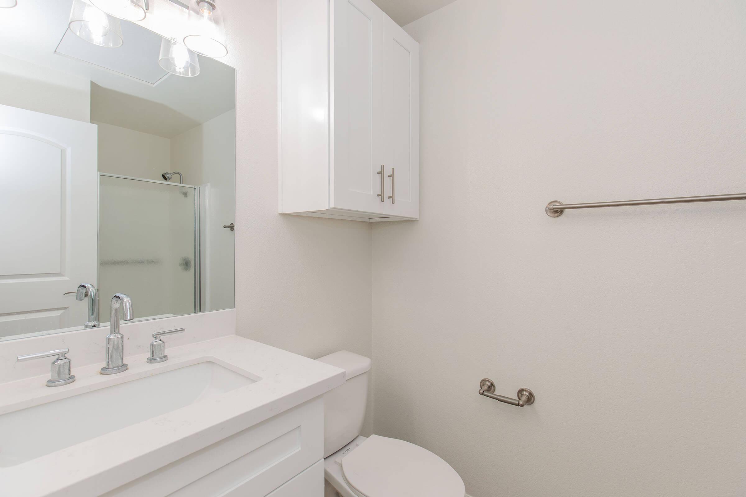 A modern bathroom featuring a white vanity with a sink, a large mirror with four light fixtures above, a shower in the background, and a white toilet. The walls are painted a light color, and there is a towel rack beside the toilet.