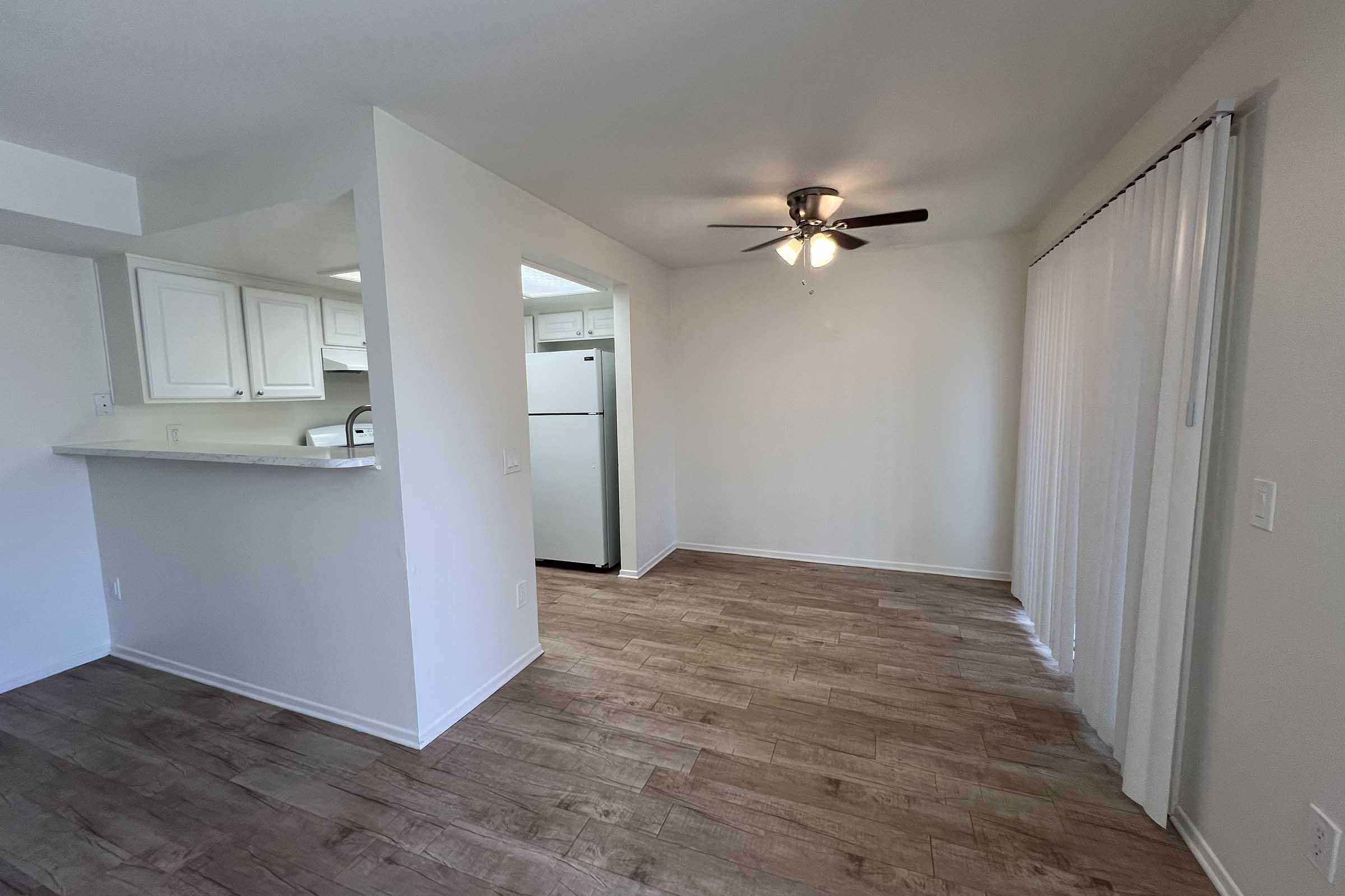 A bright, empty room featuring light brown wood-like flooring, a ceiling fan, and white walls. To the left, there is a kitchen area with a bar counter and a white refrigerator visible. Sheer curtains are positioned at a sliding door that leads outside. The overall atmosphere is clean and spacious.