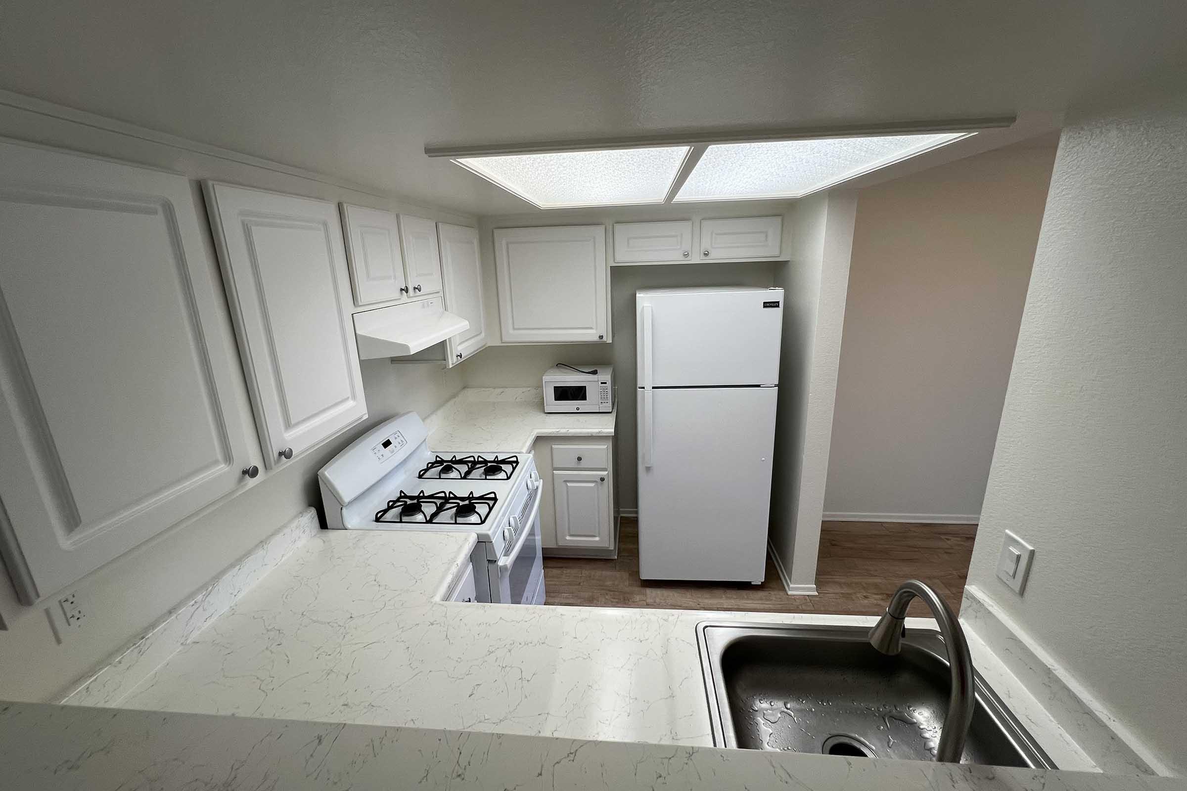 A modern kitchen view from above, featuring white cabinets, a gas stove, a microwave, and a large refrigerator. The countertops are light-colored, and a stainless steel sink is visible in the foreground. The space is well-lit with overhead lights, and the flooring appears to be wood.
