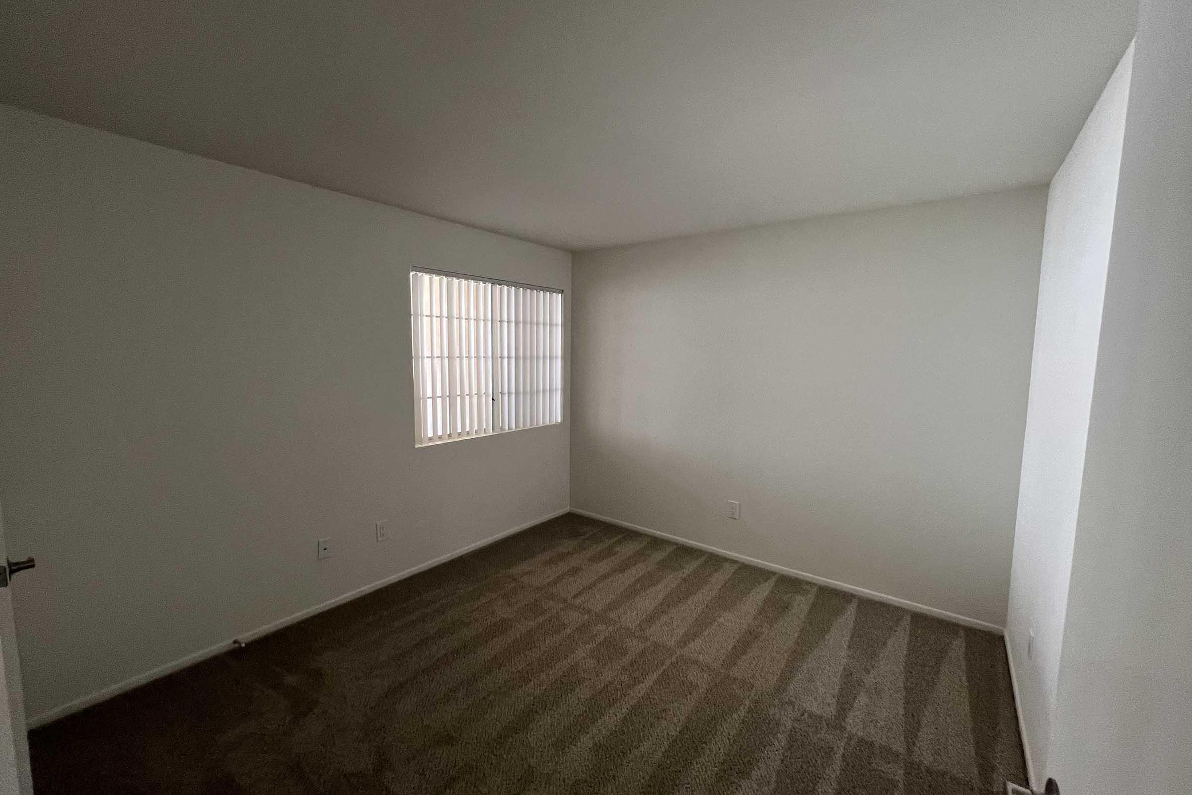 A small, empty room with plain white walls and beige carpet. There's a barred window on the left, allowing natural light to filter in. The space is unadorned and minimalistic, giving it a clean and airy feel, suitable for various uses.