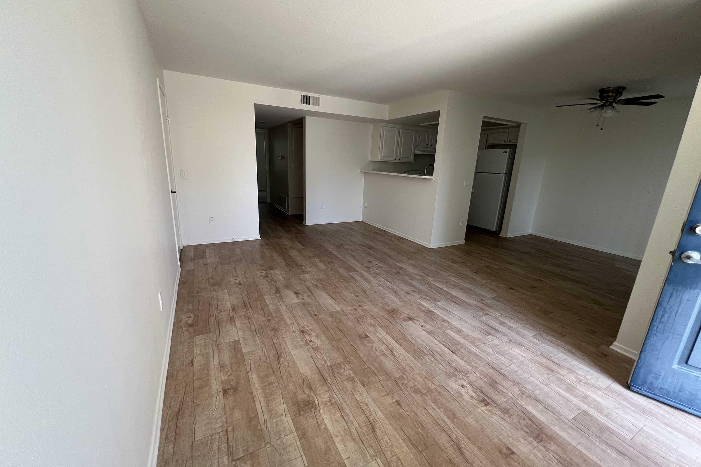 A spacious, empty living area featuring light-colored walls and wood-like flooring. To the right, a doorway leads to a kitchen with white cabinets and appliances. Natural light is coming through a nearby door, which is painted blue. The overall atmosphere is bright and clean.