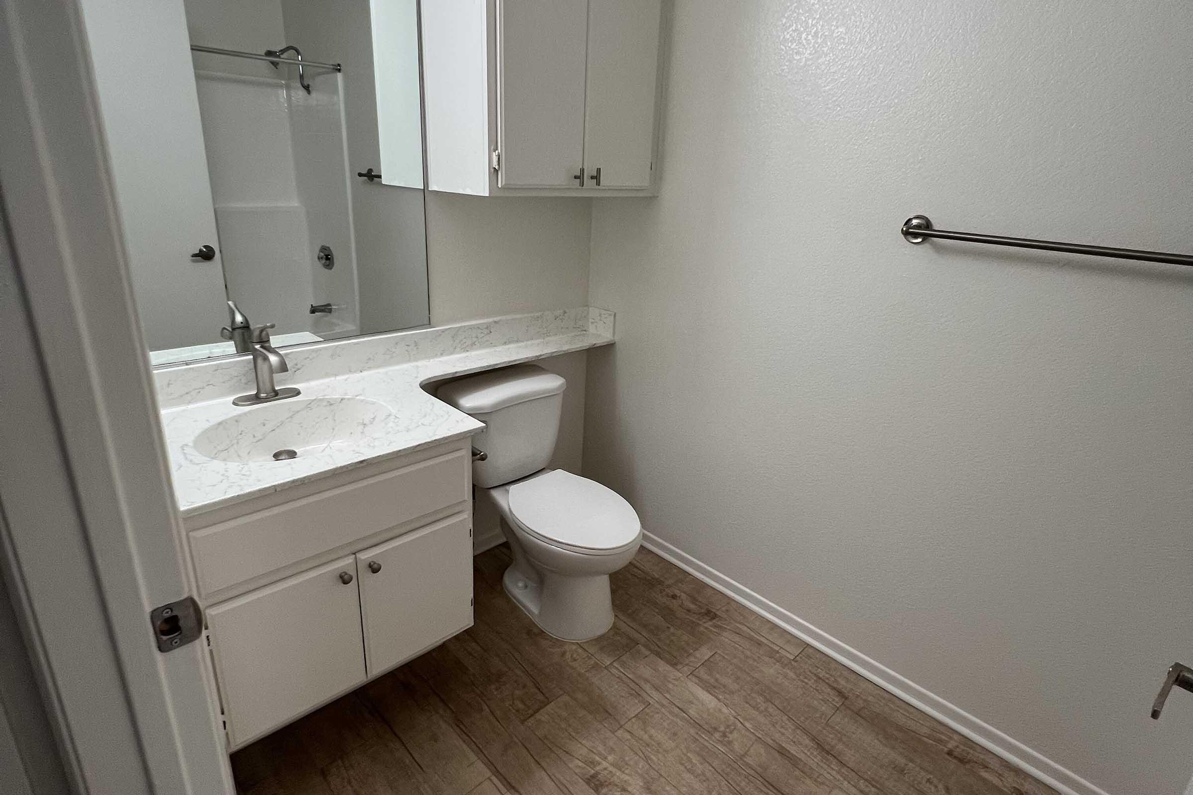 A clean, well-lit bathroom featuring a white countertop with a sink, a modern toilet, and a shower section. The walls are painted a light color, and there are white cabinets above the sink, with tiled flooring. A towel rack is mounted on the wall, contributing to the minimalist design.