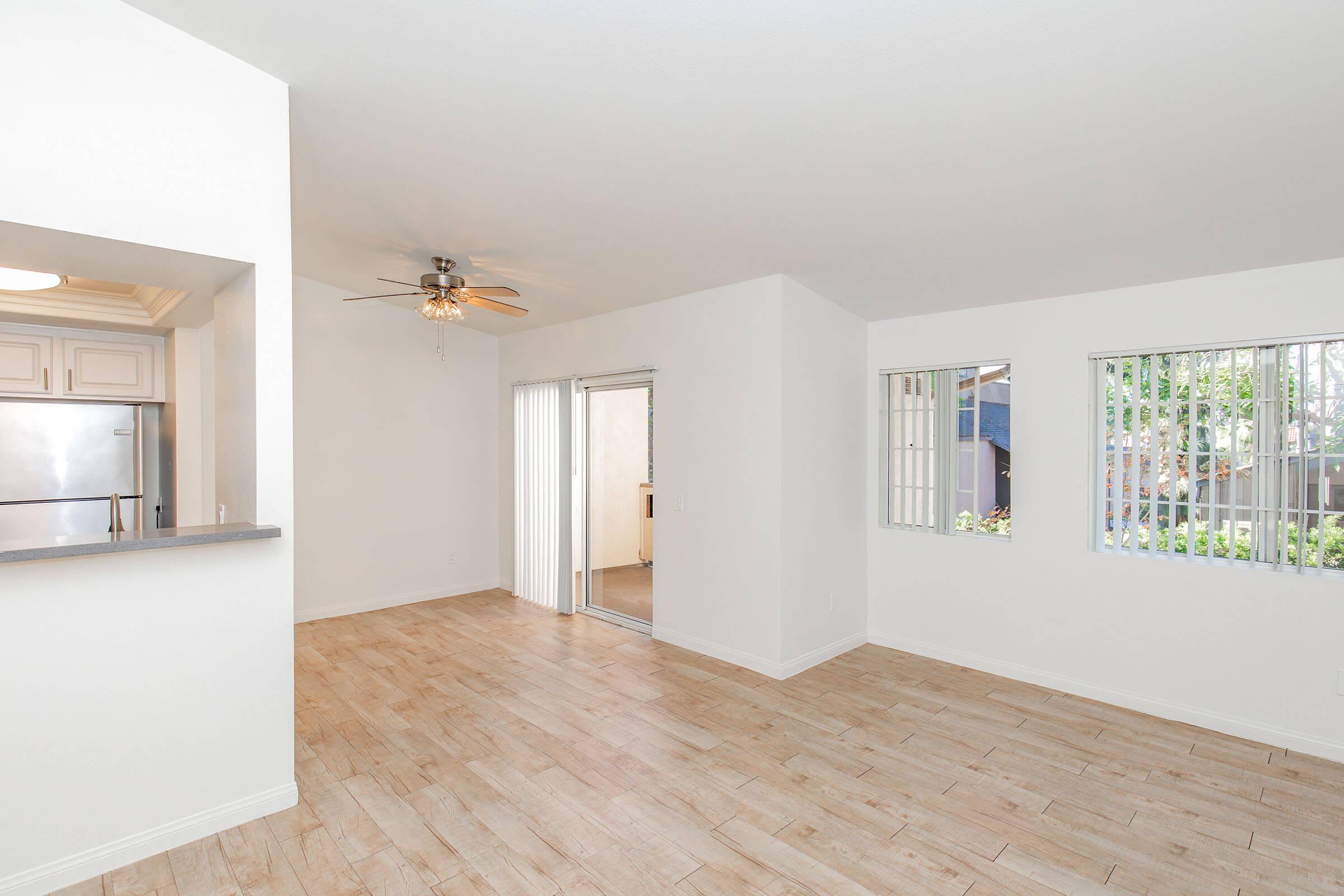 Bright and spacious living area with light-colored walls and wood-like flooring. Large windows provide natural light, and there is a ceiling fan for ventilation. A doorway leads to an outdoor area, while the kitchen area is partially visible, featuring a stainless steel refrigerator.