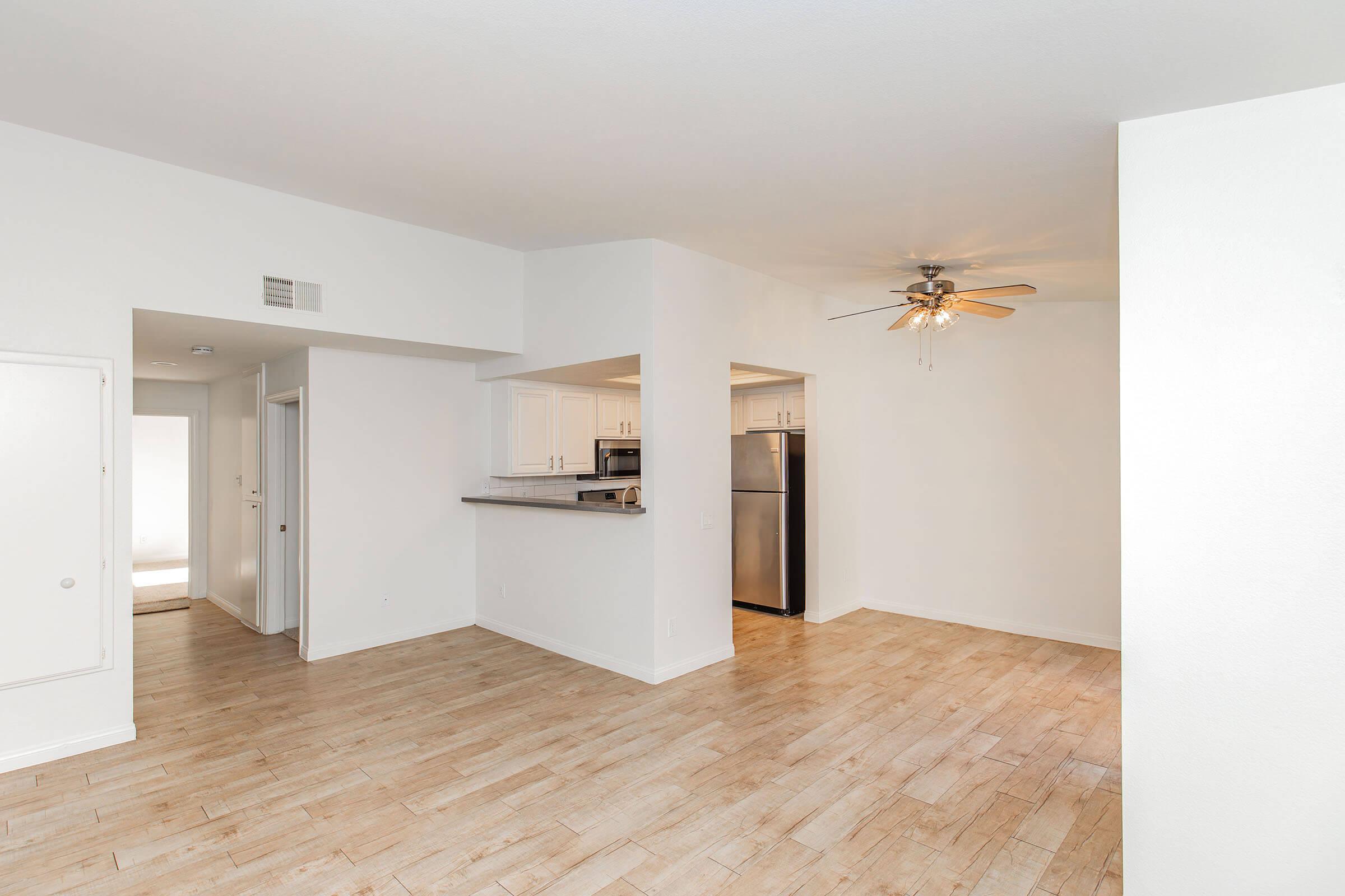 Spacious living area with light wood flooring, ceiling fan, and modern appliances. Includes a galley-style kitchen visible in the background, along with a doorway leading to another room. Walls are painted white, creating a bright and airy atmosphere.