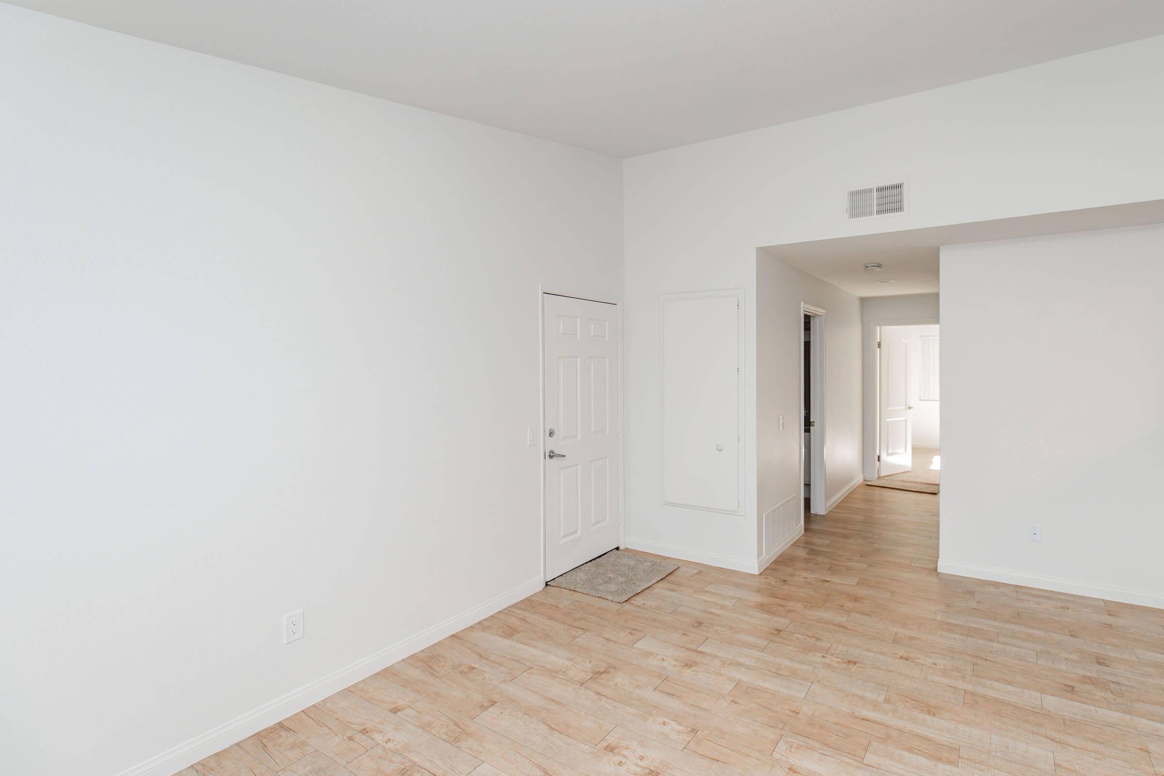 A spacious, empty room with light-colored walls and wooden flooring. There is a closed door on the left, a small welcome mat in front of it, and an entrance to another room visible in the background. The overall atmosphere is bright and clean, suggesting a newly renovated space.