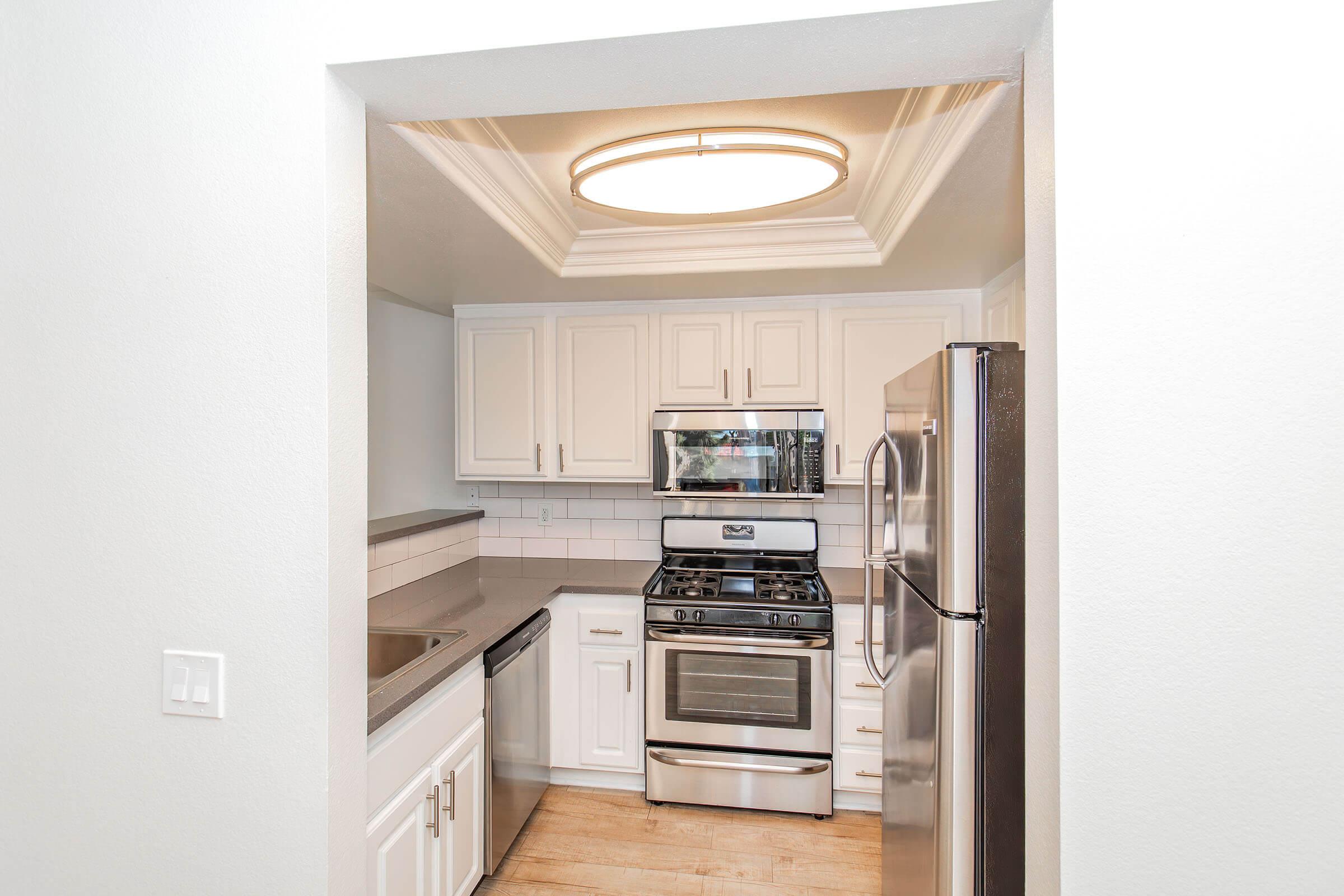 A modern kitchen featuring white cabinetry, stainless steel appliances, and a large overhead light fixture. The room includes a gas stove, microwave, and a countertop. Natural light may enter from an unseen window outside the frame, highlighting the clean and inviting space.