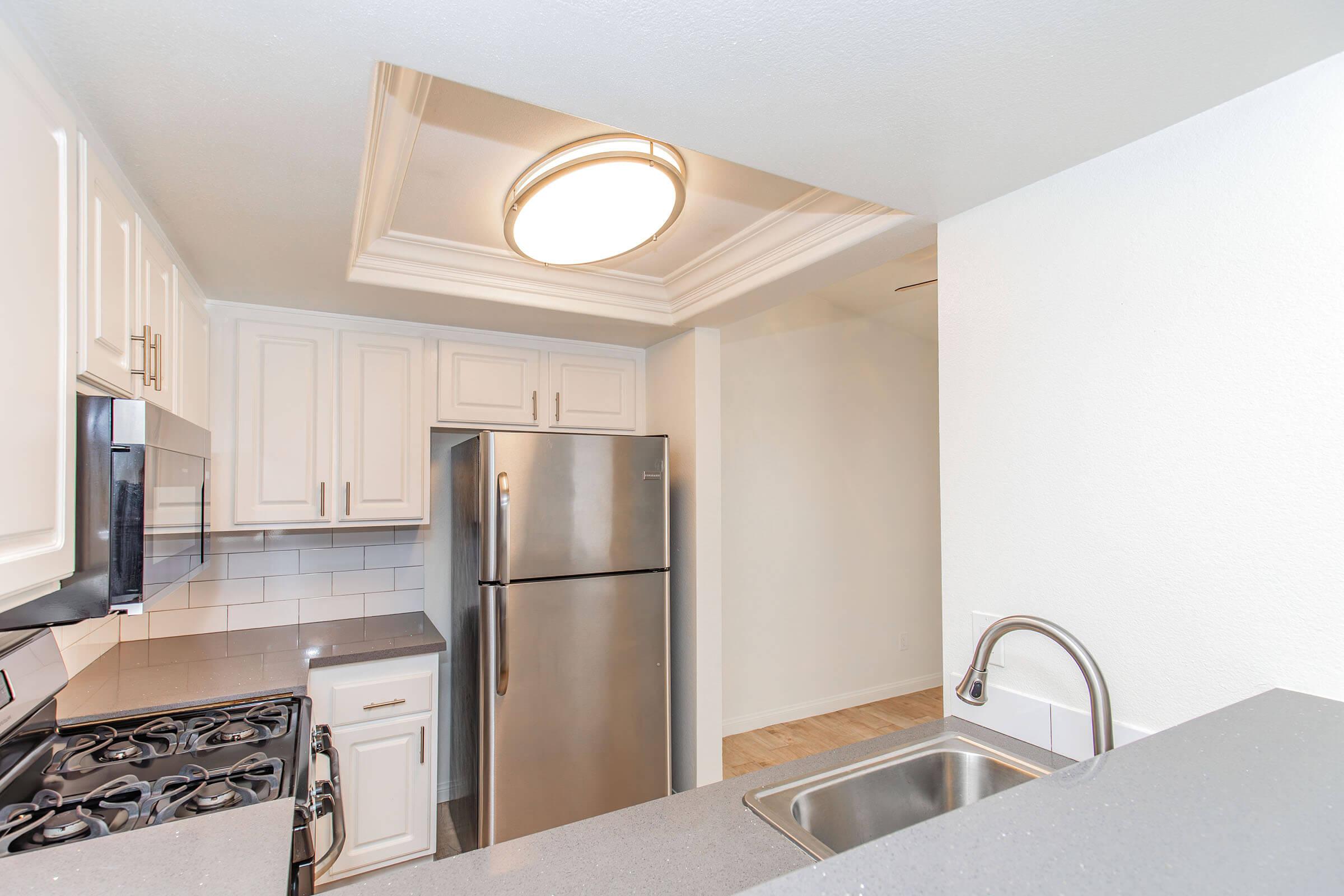 Modern kitchen featuring white cabinetry, stainless steel appliances, and a sleek countertop. Includes a gas stove, refrigerator, and a sink with a contemporary faucet. The space is well-lit with a circular ceiling light and has a clean, open layout.
