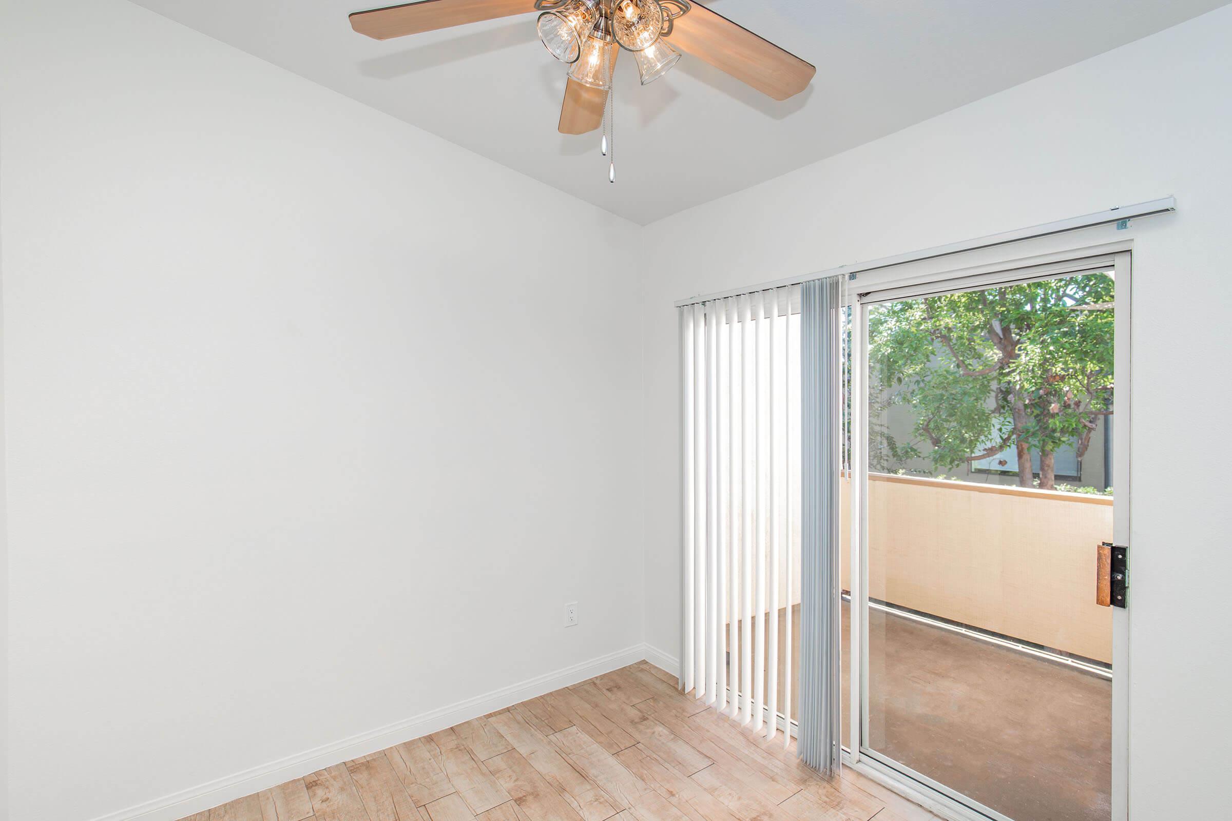 Bright, empty room with light-colored walls and wooden flooring. A ceiling fan with wooden blades is installed. Large sliding glass doors with vertical blinds lead to a small outdoor area with greenery visible outside. The room has a spacious feel and natural light.