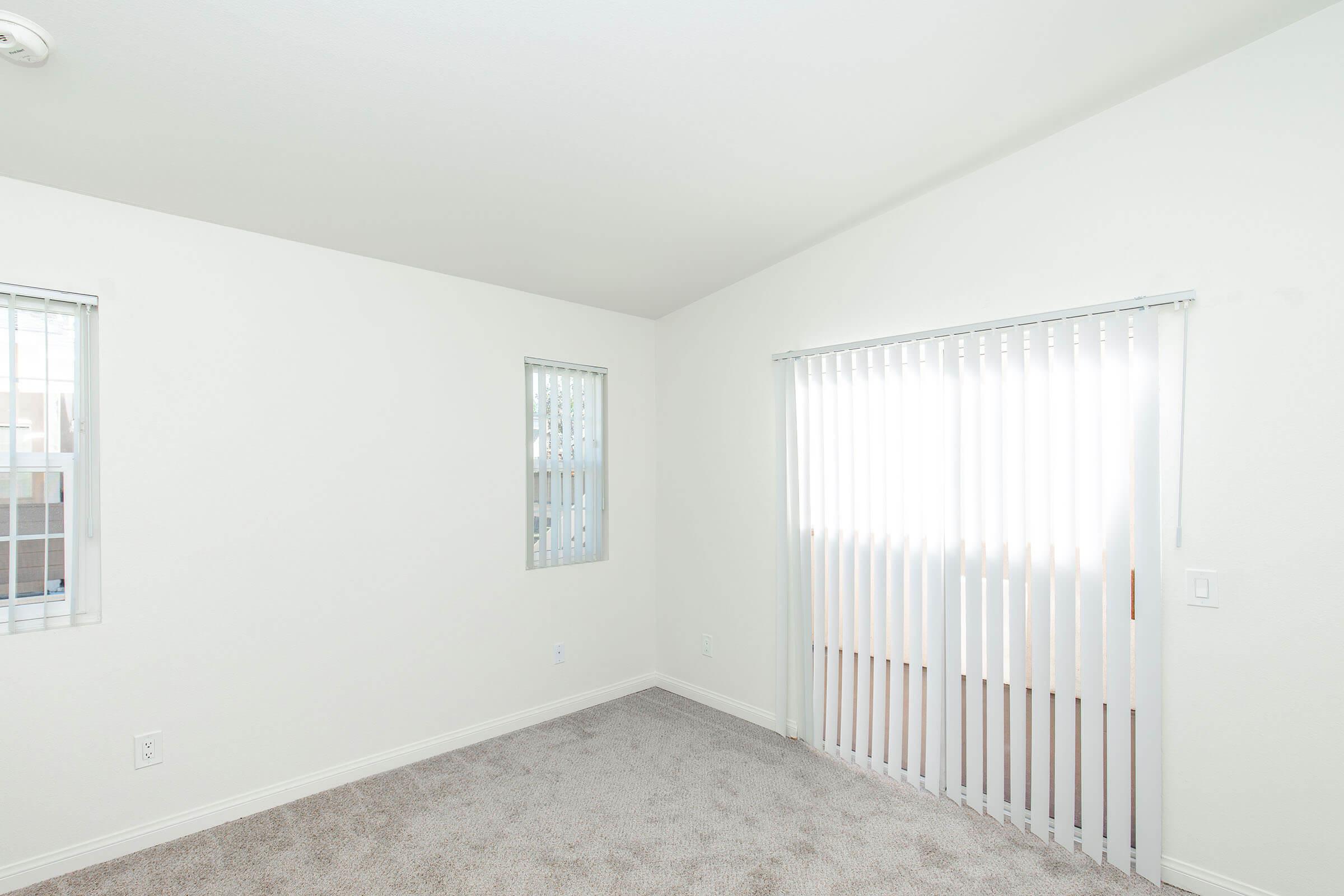 A bright, empty room with white walls and light-colored carpeting. Two windows are present: one with vertical blinds and another smaller window. The space is uncluttered, creating a minimalist ambiance.