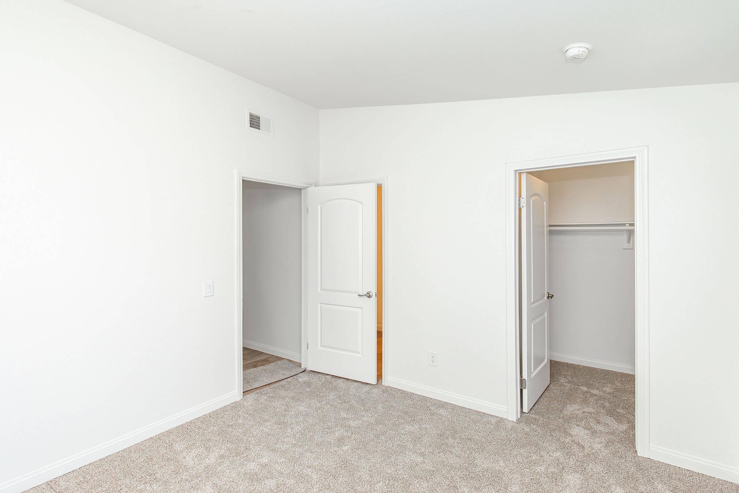 A clean, empty room with light-colored walls and carpeted flooring. There are two doors: one leading to a closet and the other likely leading to another room or hallway. The overall space is bright and uncluttered, ideal for personal decoration or furniture arrangement.