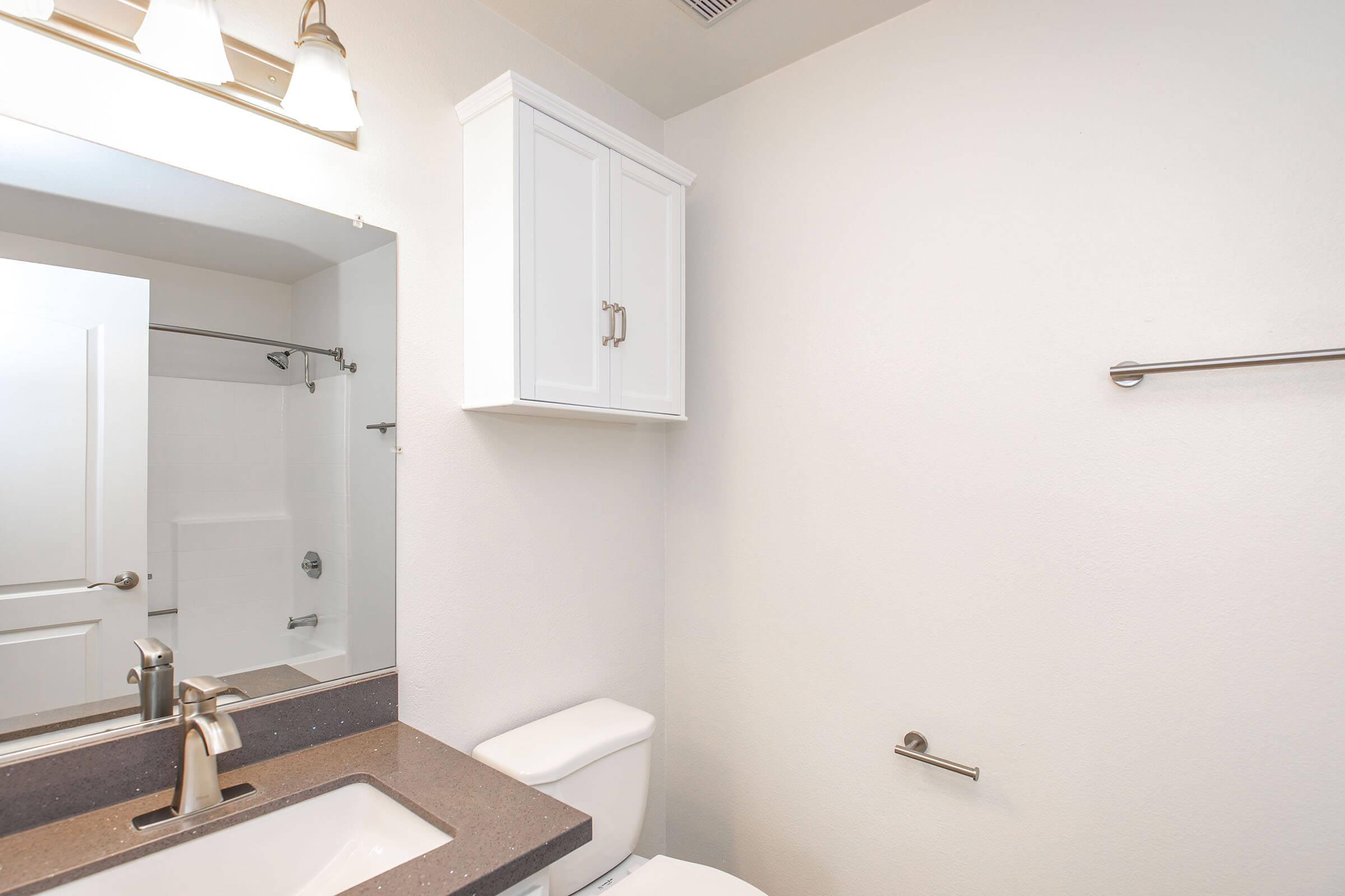 A modern bathroom featuring a large mirror above a sink with a faucet, a white toilet, and a light-colored wall. It includes a shower area with a glass door and a white cabinet mounted above the sink for storage. The overall design is clean and minimalist.