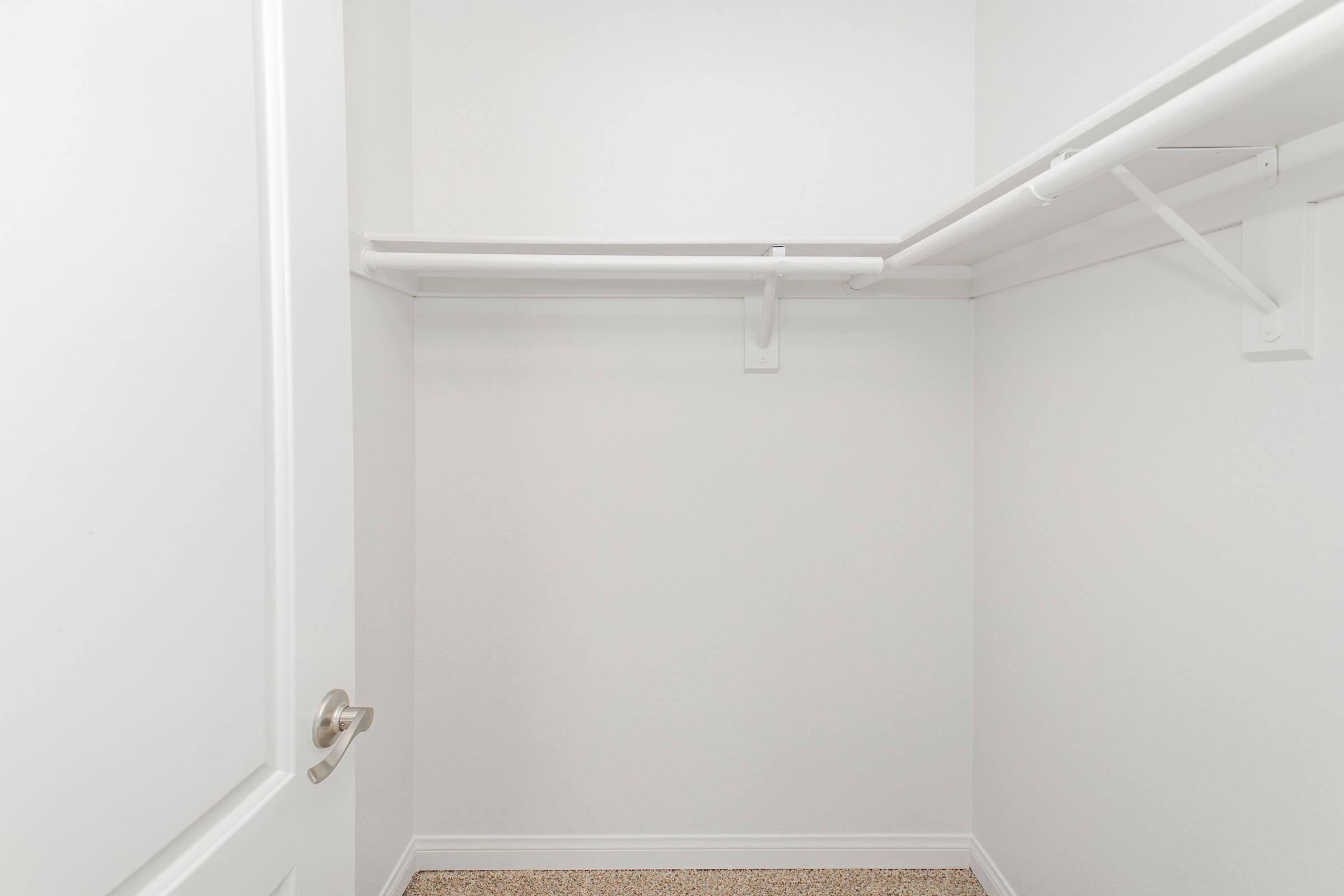An empty walk-in closet with white walls and a carpeted floor. There are two hanging rods installed on the walls, providing space for clothing storage. A closed door is visible on the left side of the image. The overall aesthetic is minimalistic and clean.