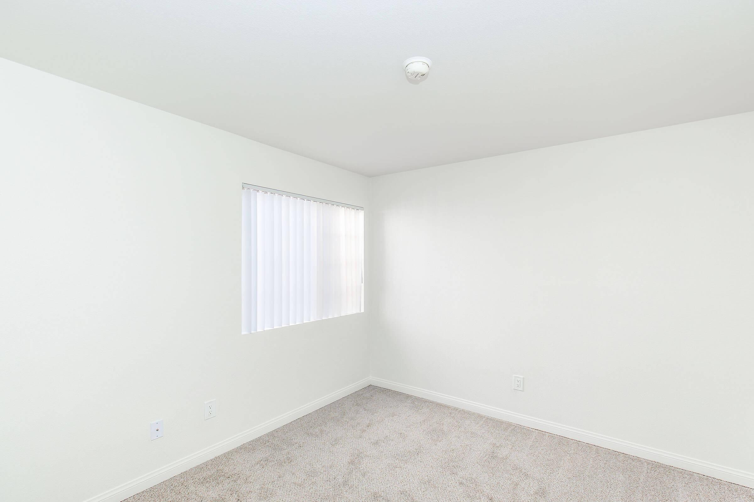 A minimalistic interior of an empty room with light gray carpet, white walls, and a window with vertical blinds, allowing natural light to enter. The ceiling features a single light fixture, and there are no furnishings or decorations visible, creating a spacious and clean environment.