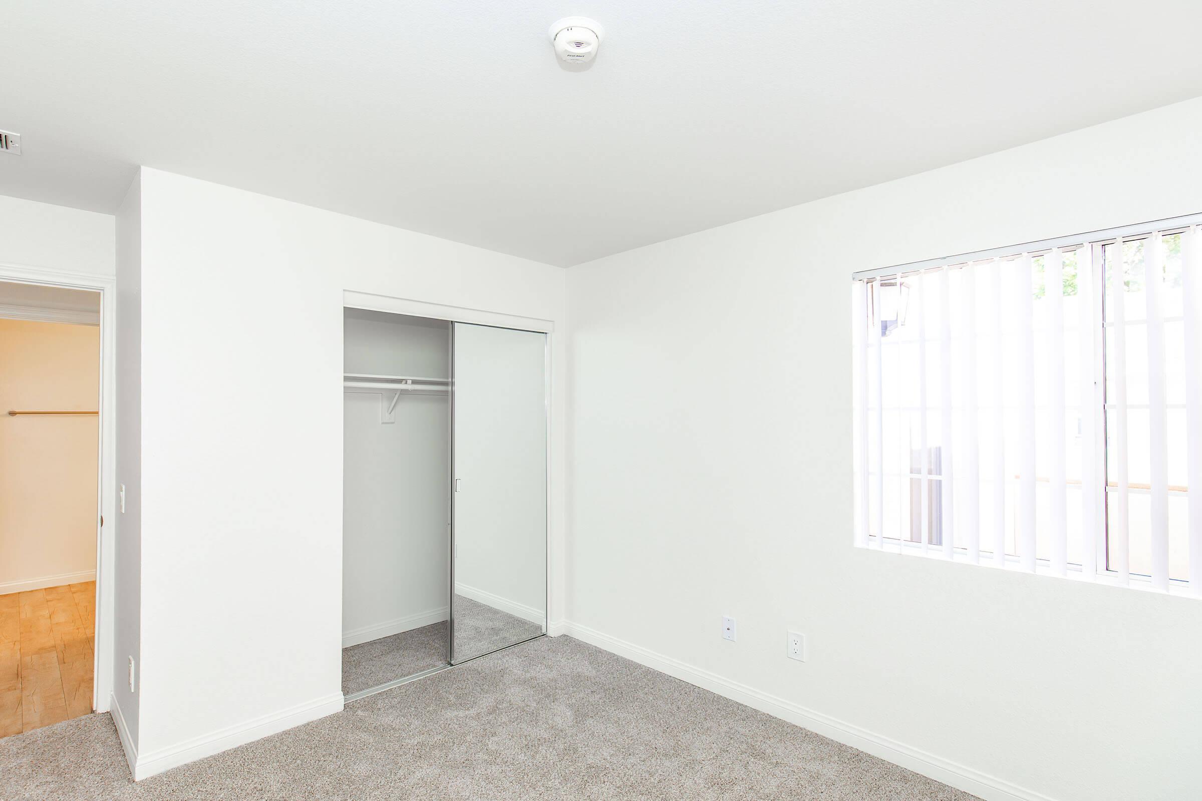 A vacant bedroom featuring light gray carpet, white walls, and a mirrored closet. Natural light enters through a window with vertical blinds. The room has simple decor and a clean, modern aesthetic, creating an open and airy feel. An additional doorway leads to an unseen area.