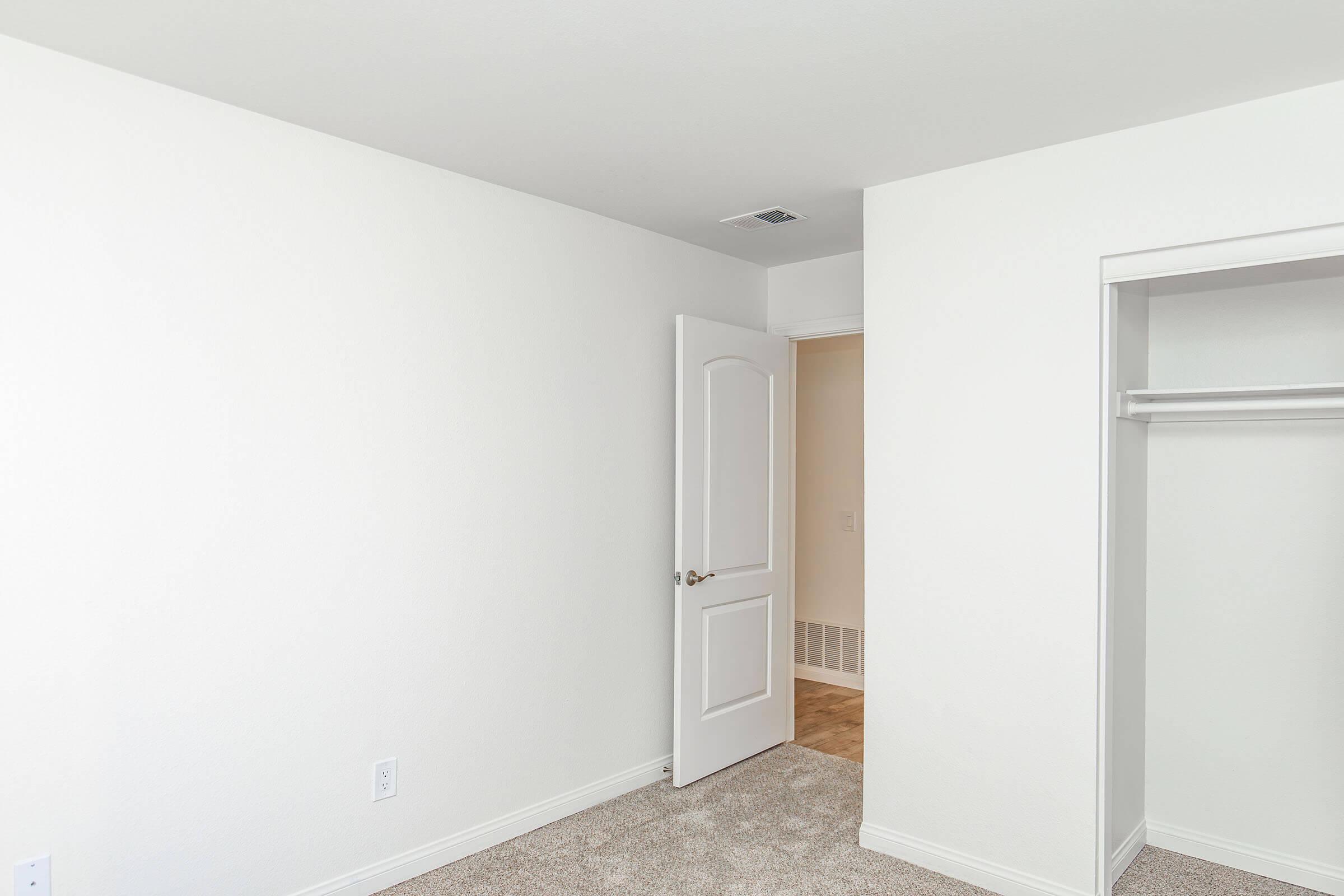 A light-colored room with beige carpeting, featuring a white door partially open to another space. The room has plain white walls and a closet with no doors on the left side, showcasing a simple and clean aesthetic.