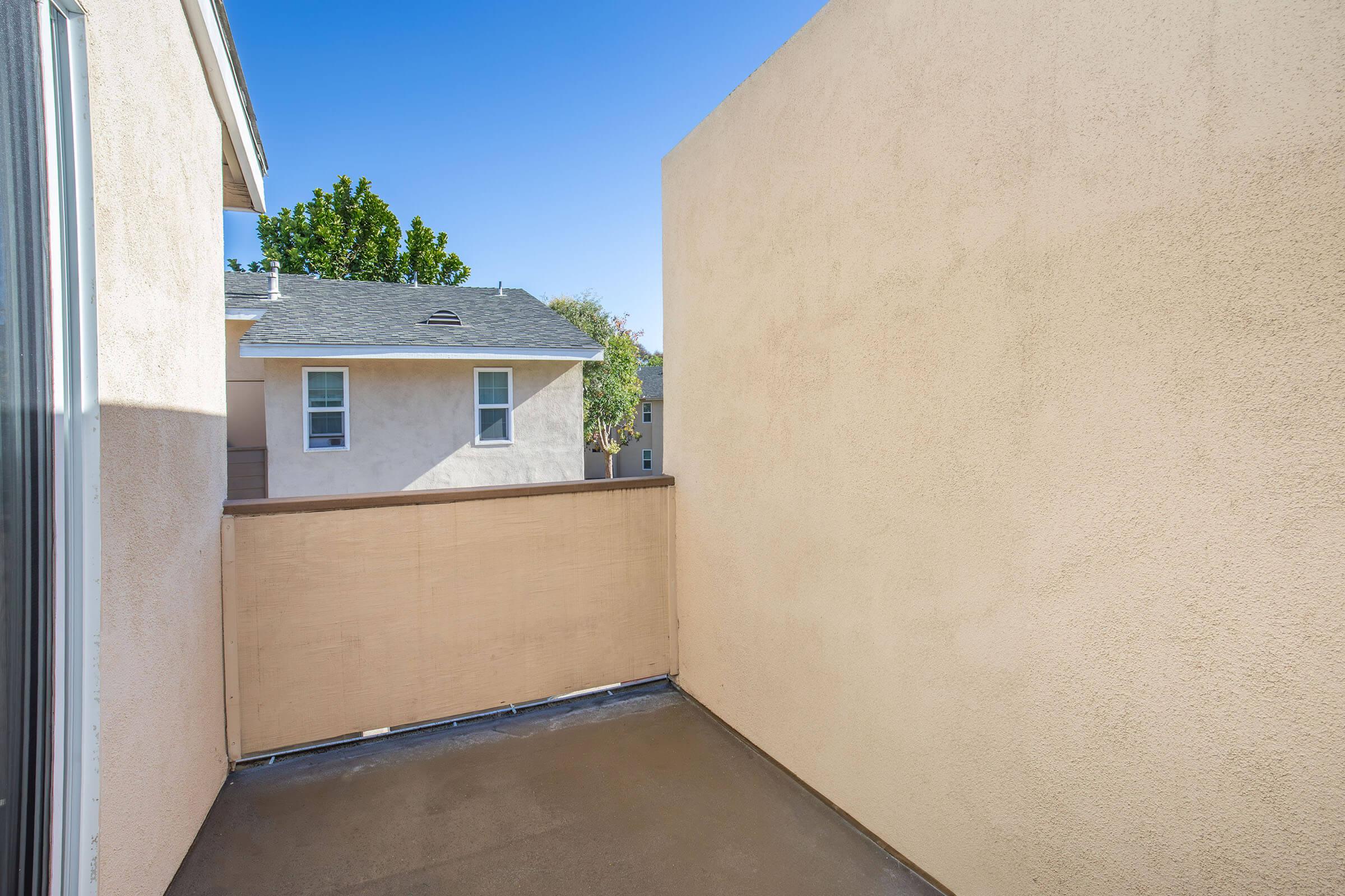 A small balcony or terrace with a beige wall, overlooking a neighborhood. The sky is clear and blue, and there are houses visible in the background. The area appears sunny and well-lit.