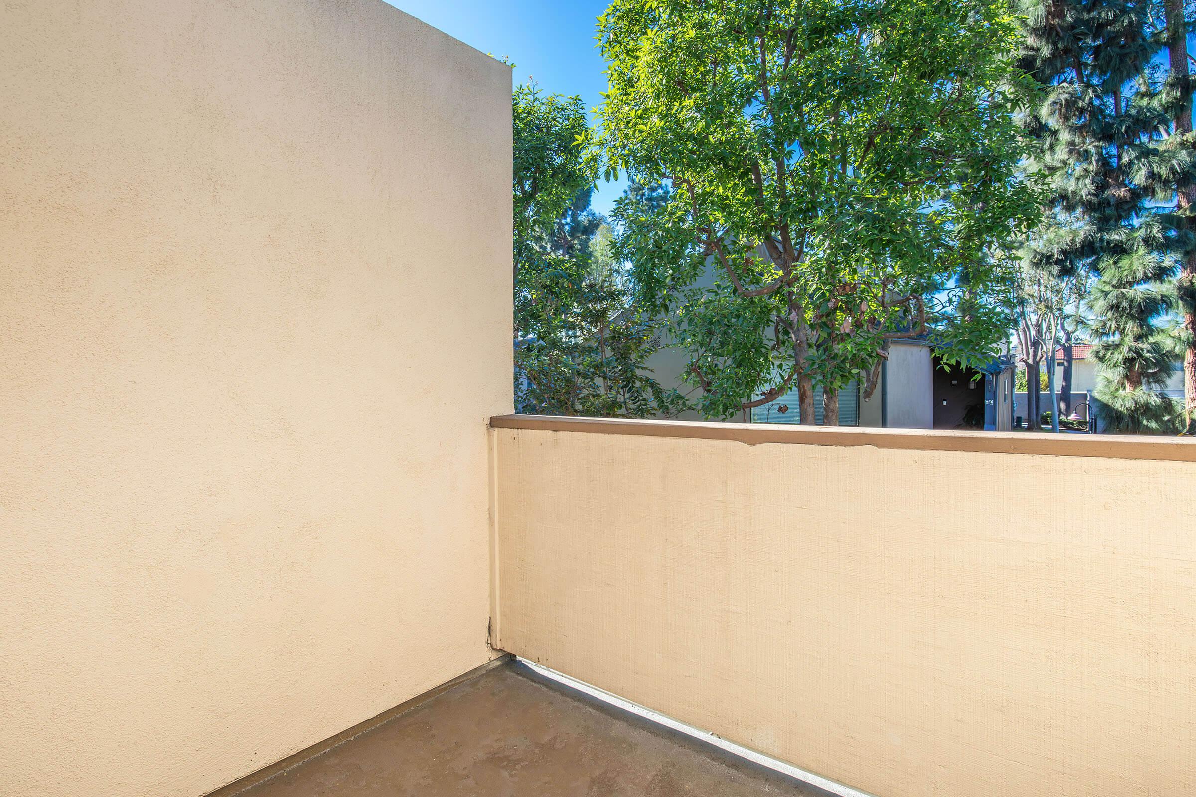 A corner balcony with beige walls and a concrete floor, surrounded by greenery. Sunlight filters through the trees, creating a bright atmosphere. The view includes neighboring buildings partially obscured by foliage.