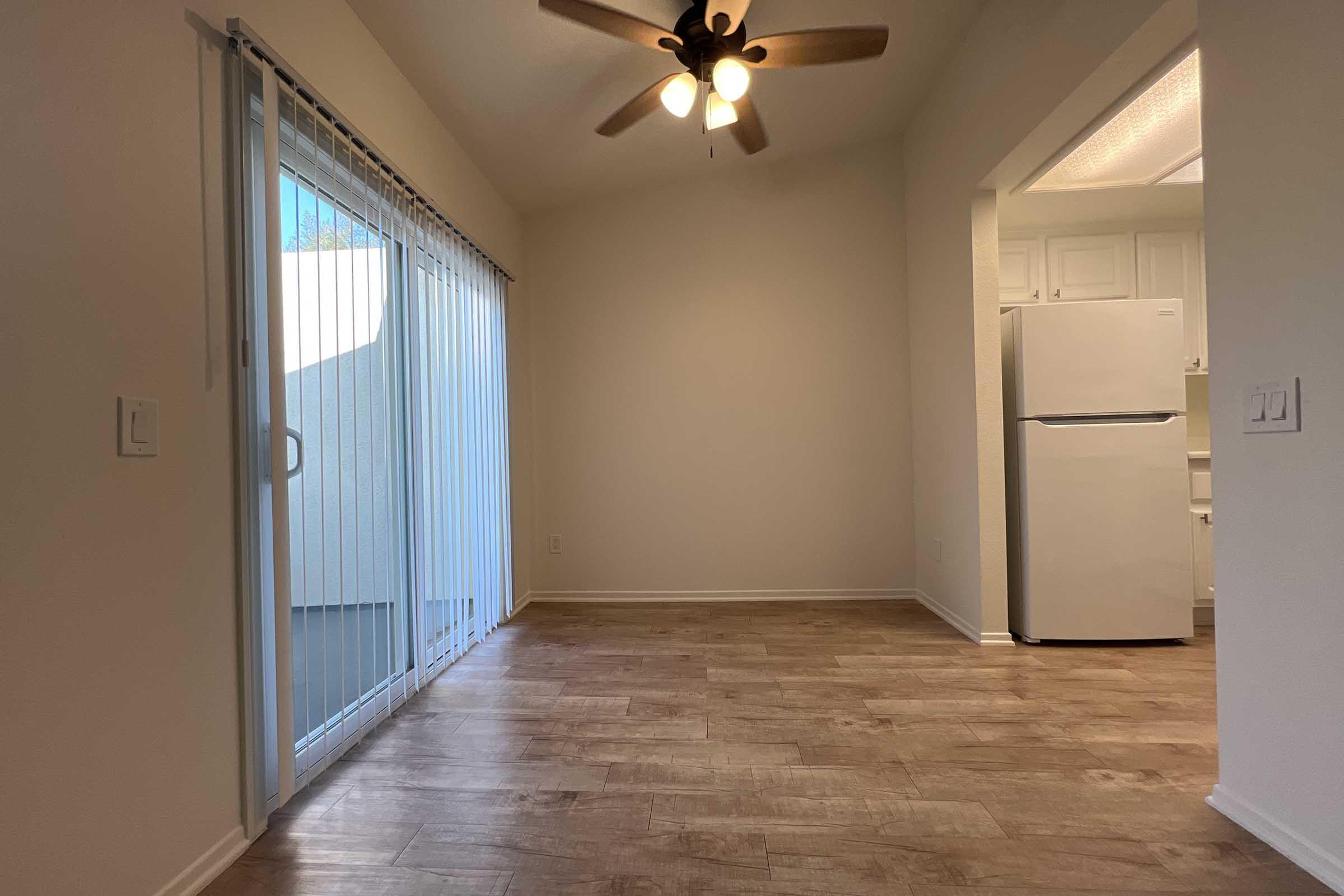 An empty room with wooden flooring, a ceiling fan, and a large sliding glass door leading to a balcony. A white refrigerator is visible in the corner, and the walls are painted a light color, creating a bright and airy atmosphere.