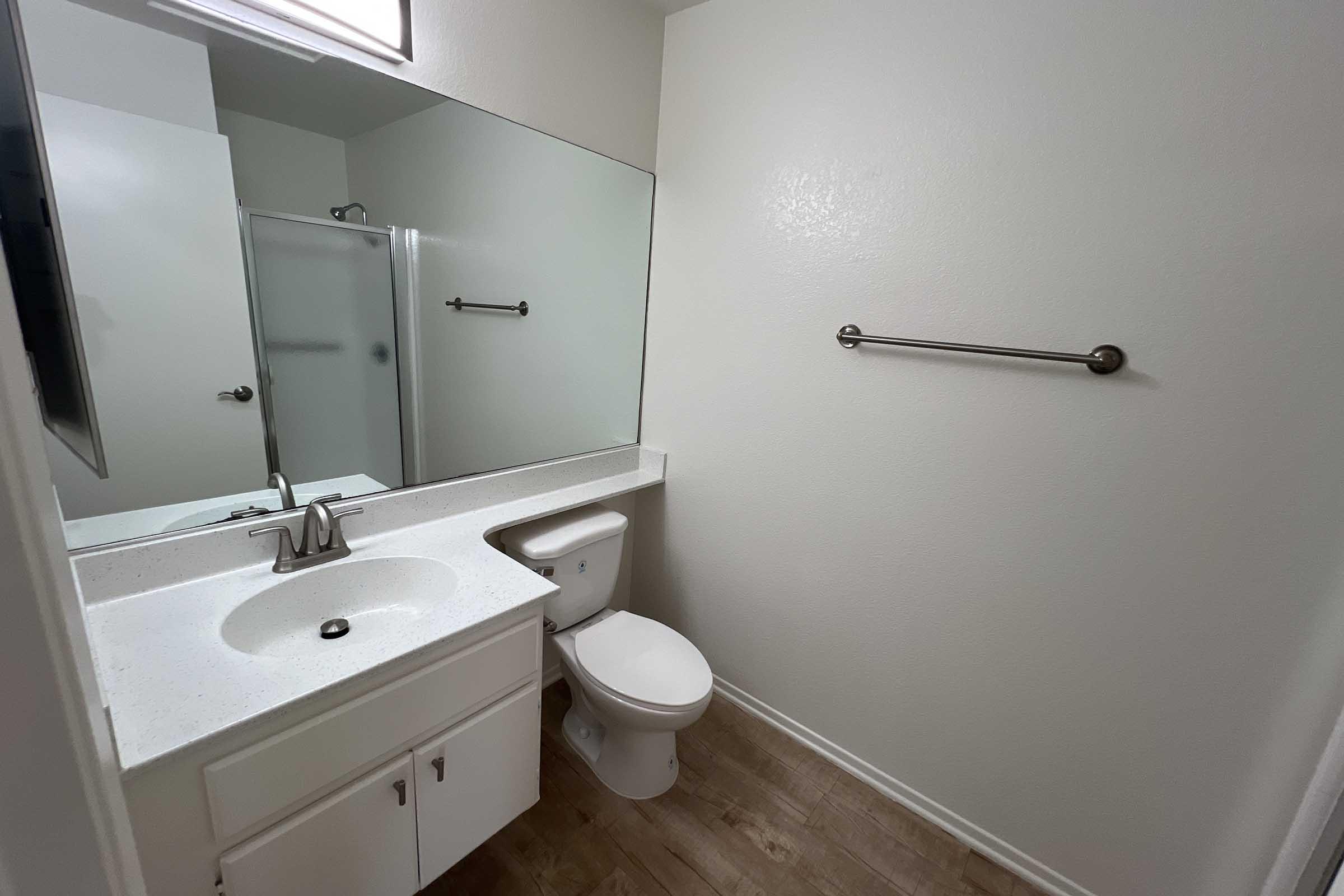 A clean, modern bathroom featuring a light-colored vanity with a sink, a toilet, and a large mirror. The walls are painted white, and there is a towel bar on one side. A glass shower is visible in the background, with wooden-style flooring throughout the space. Natural light enters from a window above the mirror.