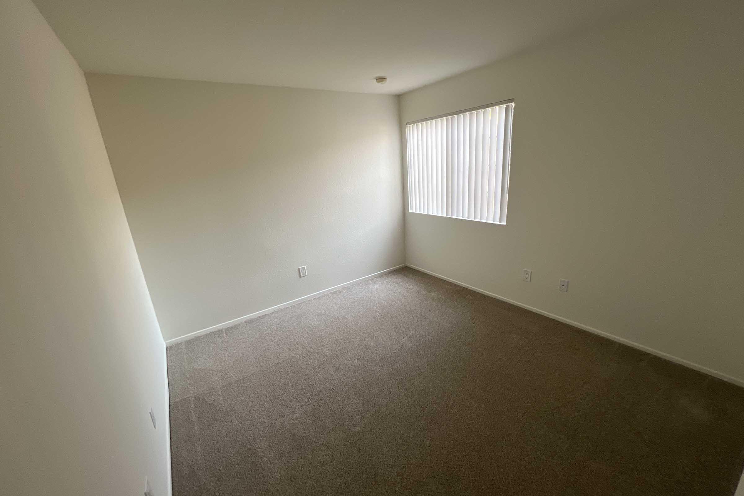 An empty room with light-colored walls and carpeted flooring. A window with vertical blinds allows natural light to enter, creating a bright atmosphere. The space is free of furniture, showcasing the room's dimensions and potential for use.