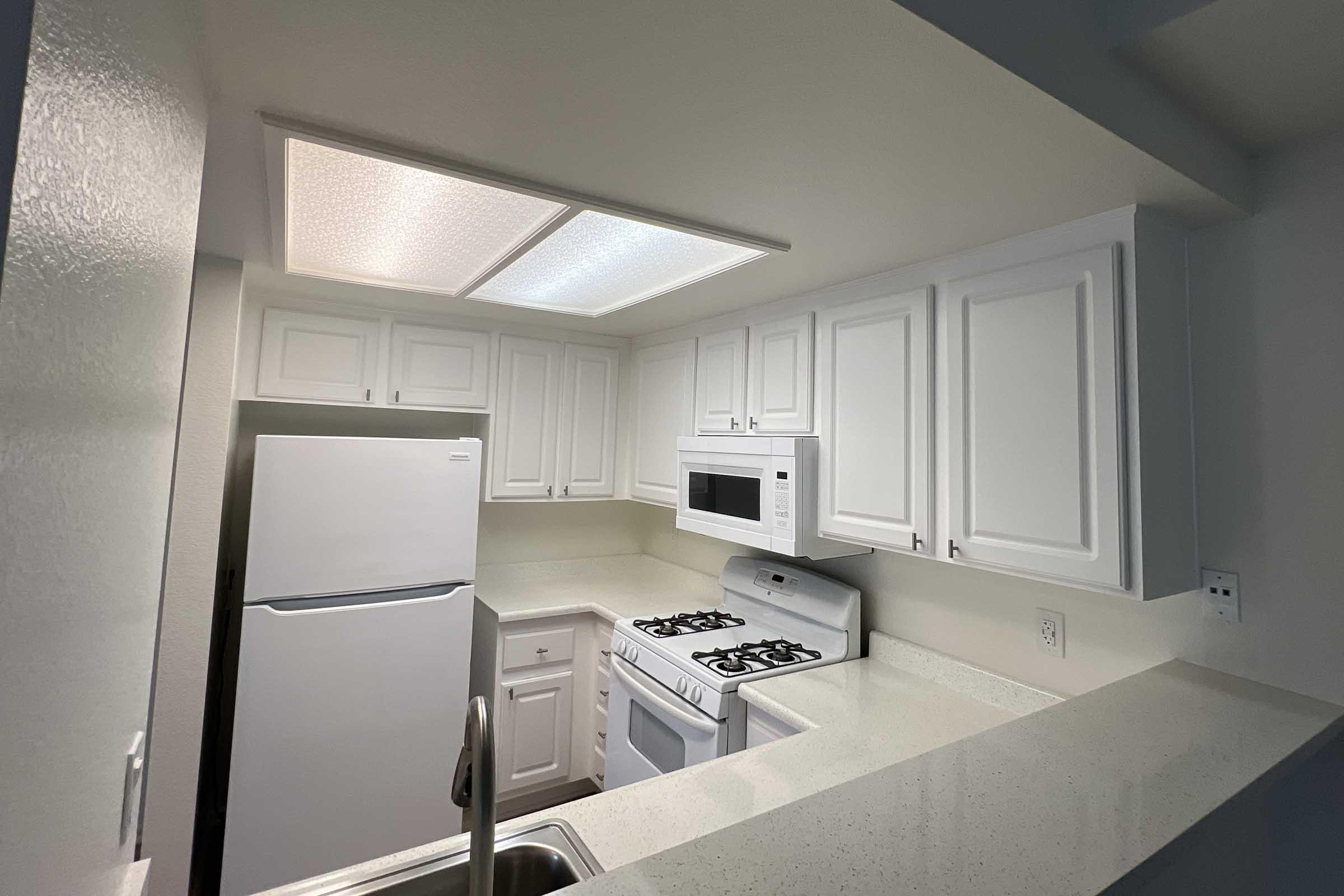 A clean, modern kitchen featuring white cabinets, a white refrigerator, a microwave above the stove, and a gas range. The countertop is light-colored, and the space is well-lit with ceiling lights. The kitchen has a sleek and inviting appearance, suitable for cooking and dining.